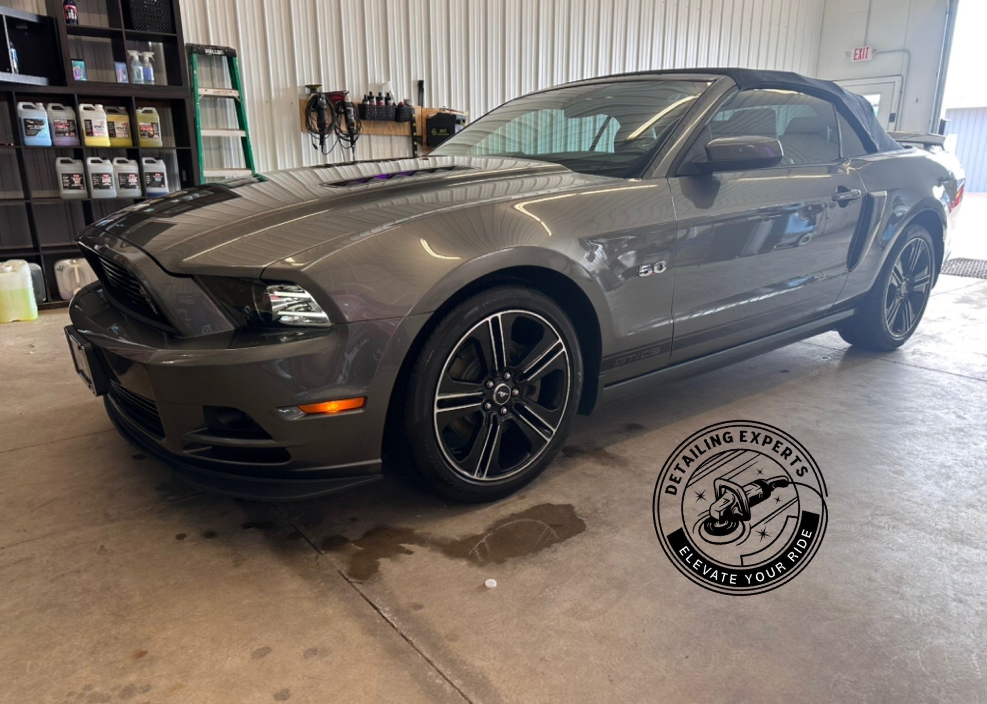 Gray Ford Mustang convertible inside a garage with shelves of car supplies, a ladder, and a logo of detailing experts on the floor.