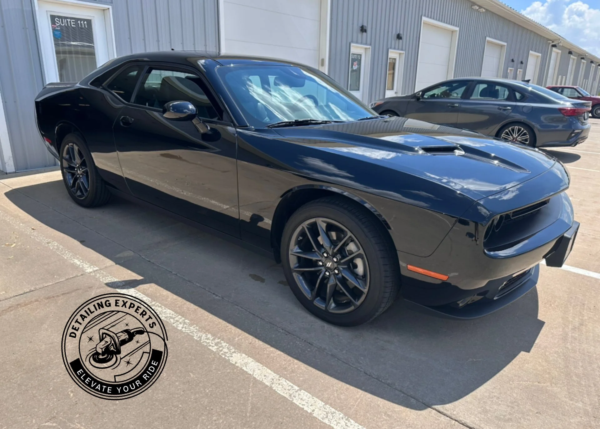 Black sports car parked in a parking lot outside a building with multiple doors and windows, and a logo in the lower left corner that reads 'Detailing Experts' with a tire and sparkles.