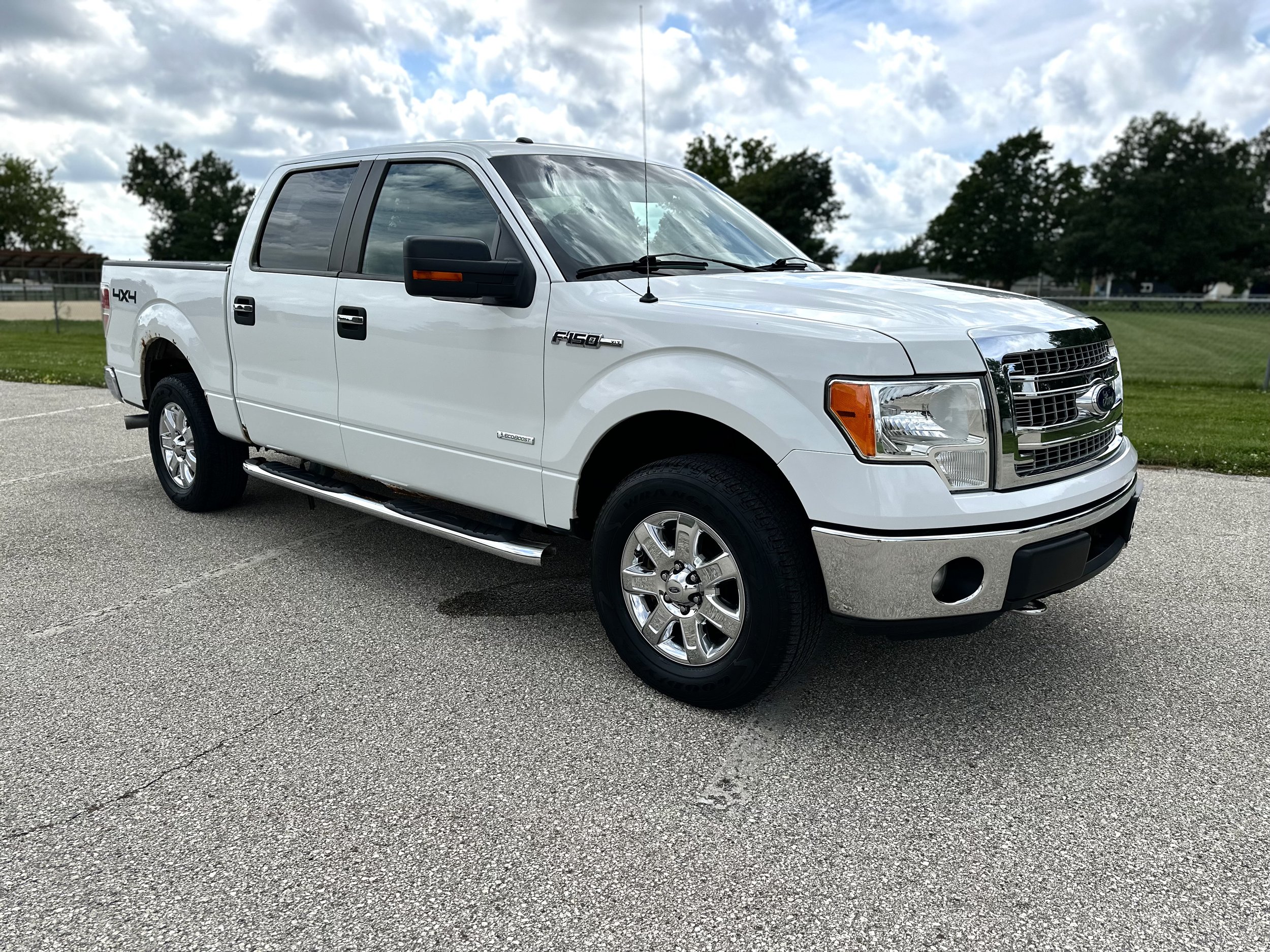 White Ford F-150 pickup truck parked on pavement with green grass and trees in the background, under a partly cloudy sky.