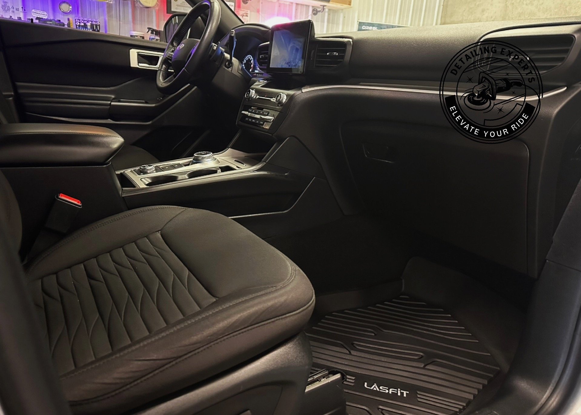 Interior of a modern vehicle showing the driver's seat, dashboard, steering wheel, and center console with a touchscreen display. The vehicle has black leather seating and rubber floor mats with 'LASFIT' branding.