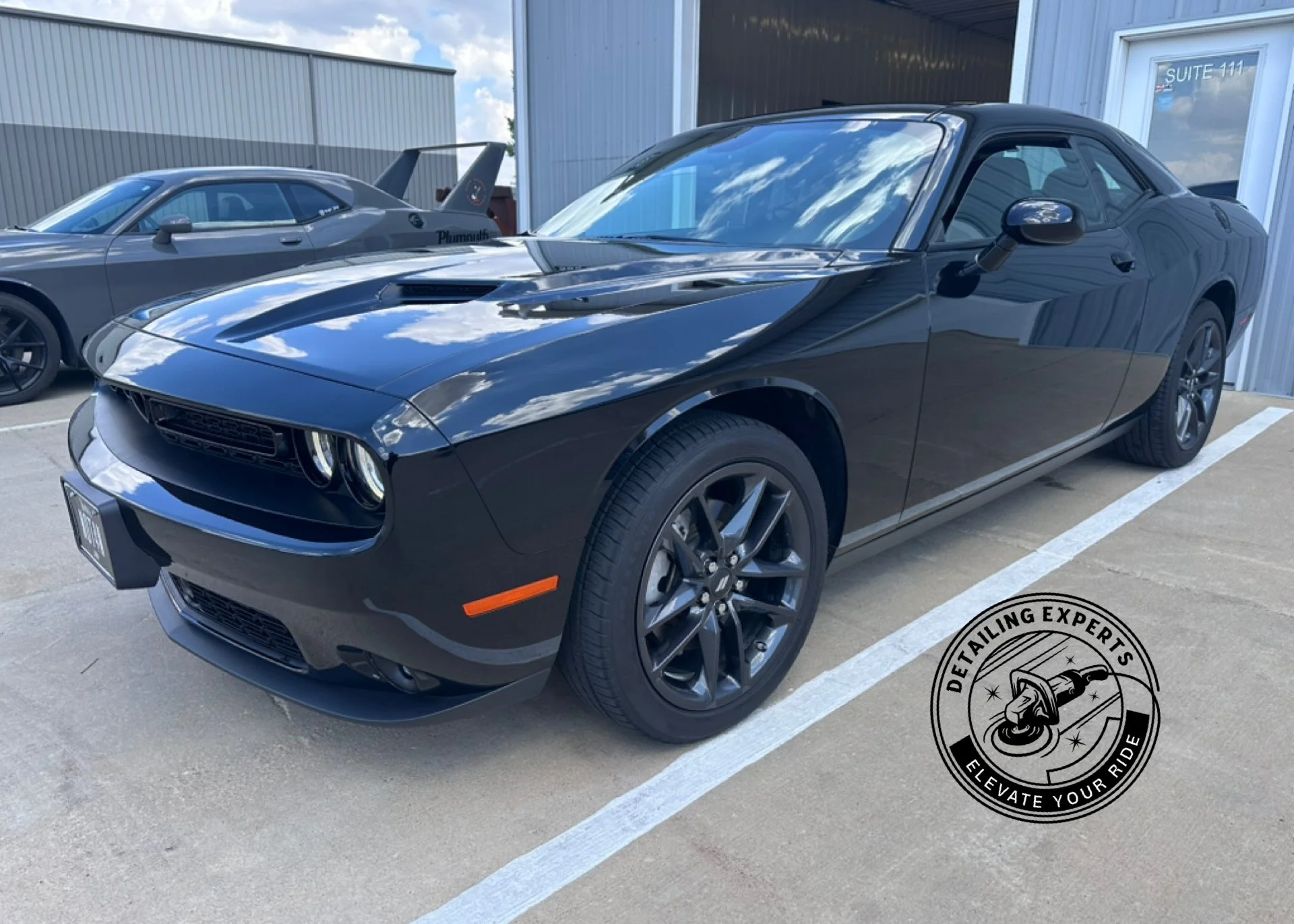 A black Dodge Challenger parked next to a grey coupe at a car dealership with a logo for detailing experts 'Elevate Your Ride' on the ground.