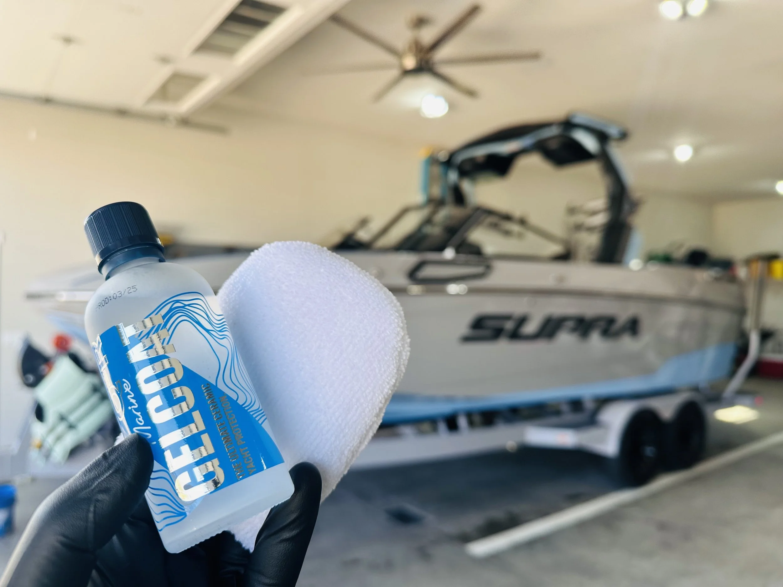 A hand wearing a black glove holding a bottle of Meguiar's Marine Rinse with a white microfiber towel in front of a boat on a trailer inside a garage.