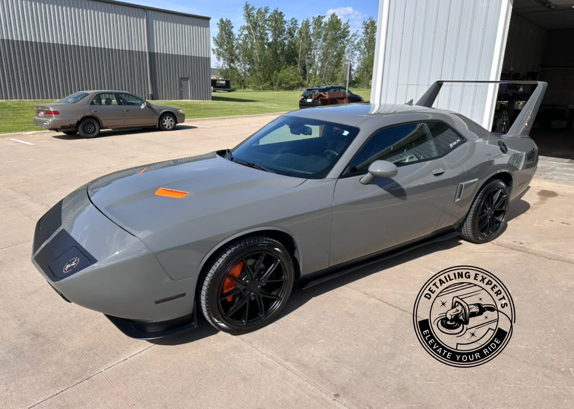 A gray Chevrolet Corvette sports car with black wheels parked outside a garage. In the background, there is a brown car and a building with trees behind it. The Corvette has a large rear wing and the logo of a detailing service with the text 'Detaili