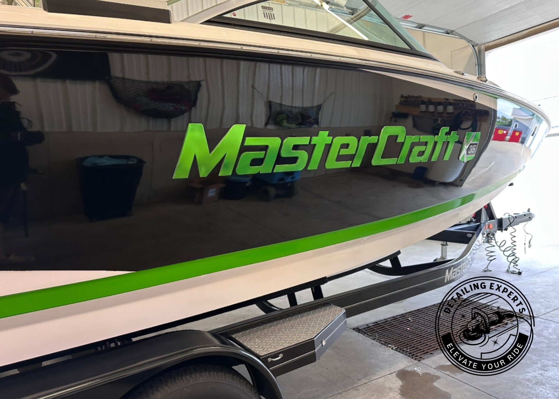 A boat on a trailer inside a garage with a black reflective side featuring the green MasterCraft logo. Part of a circular black and white logo with the text 'Detailing Experts' and 'Elevate Your Ride' is visible at the bottom right.