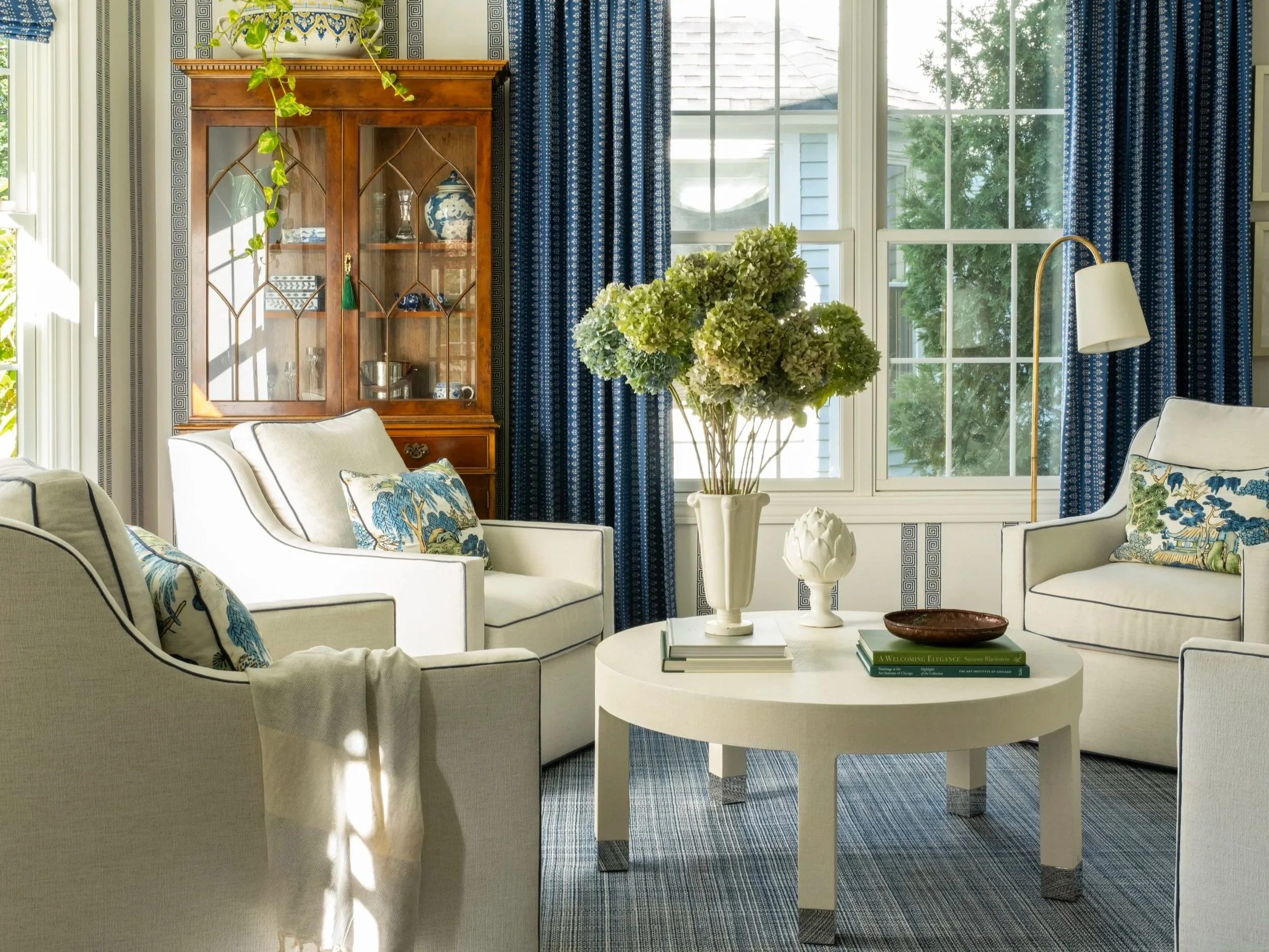 A bright living room with white seating, a central white coffee table, blue patterned curtains, a wooden display cabinet, a large vase with green hydrangeas, and decorative pillows.