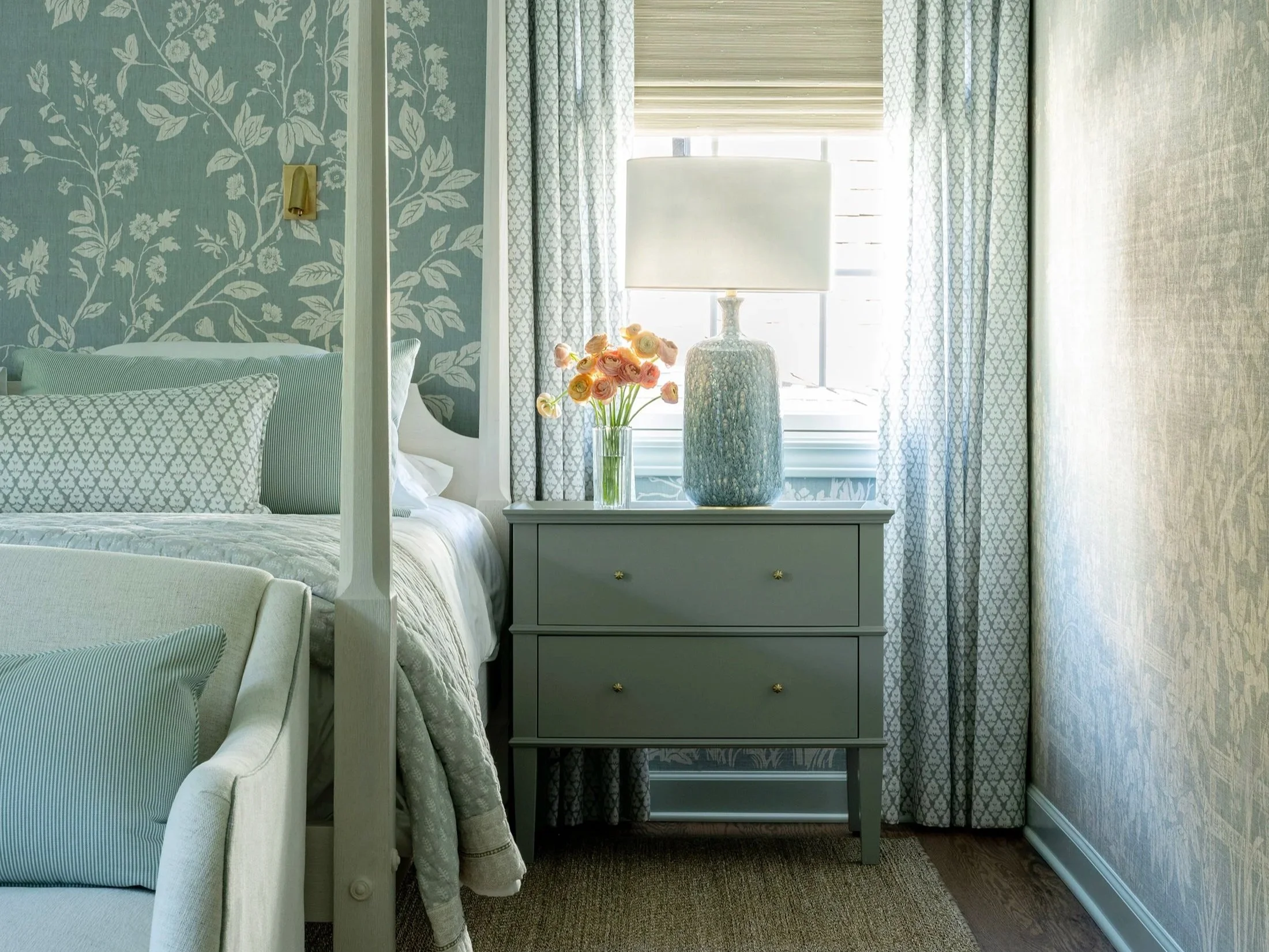 A bedroom with a bed, decorative pillows, a nightstand with a large vase, a lamp, and a vase of flowers, with patterned curtains and wallpaper.
