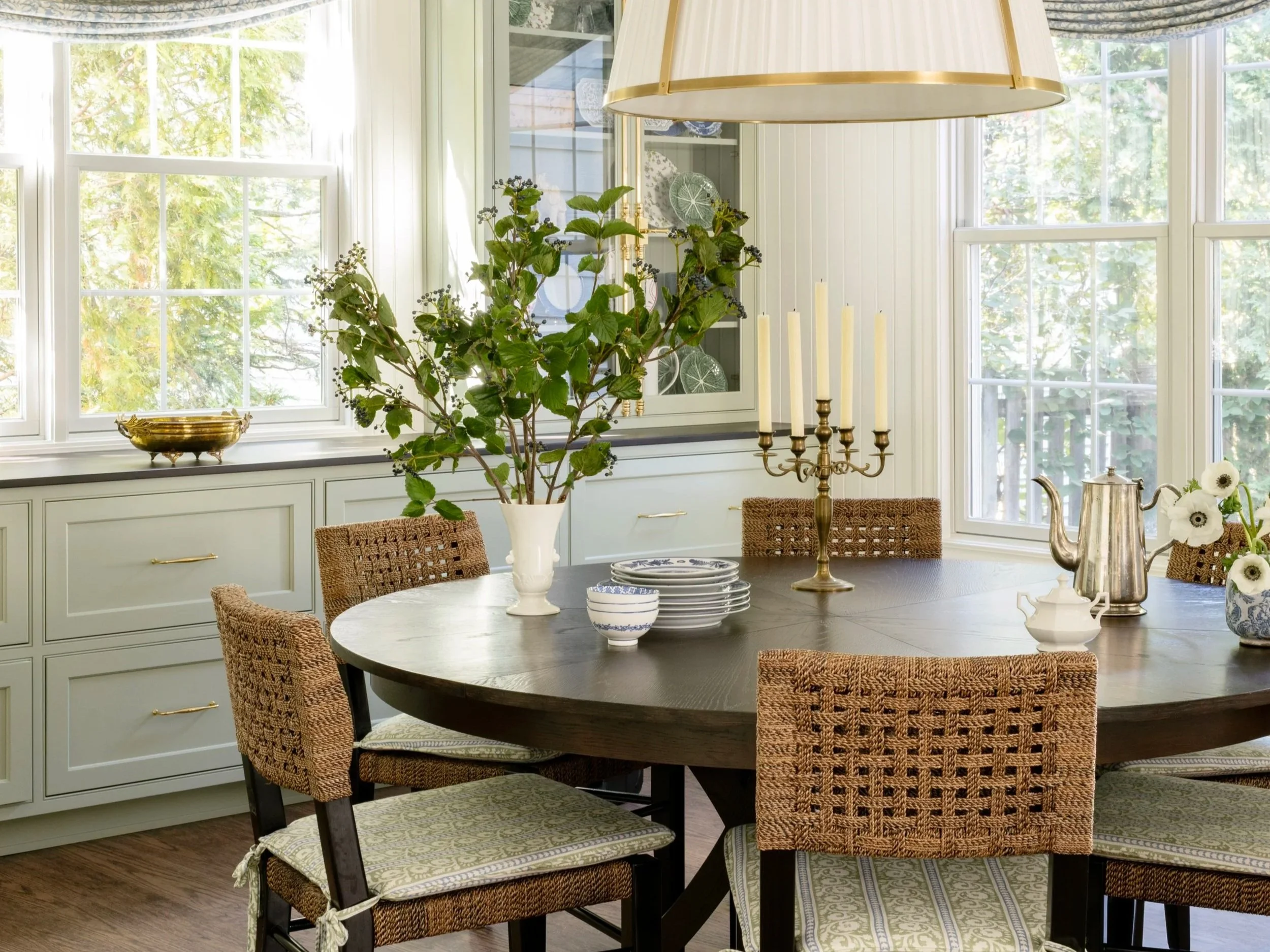 A dining room with a round wooden table, wicker chairs with cushions, and a large window that lets in natural light. The table has a white vase with green foliage, a brass candelabra, and various dishes and pitchers. Light-colored cabinetry with gold