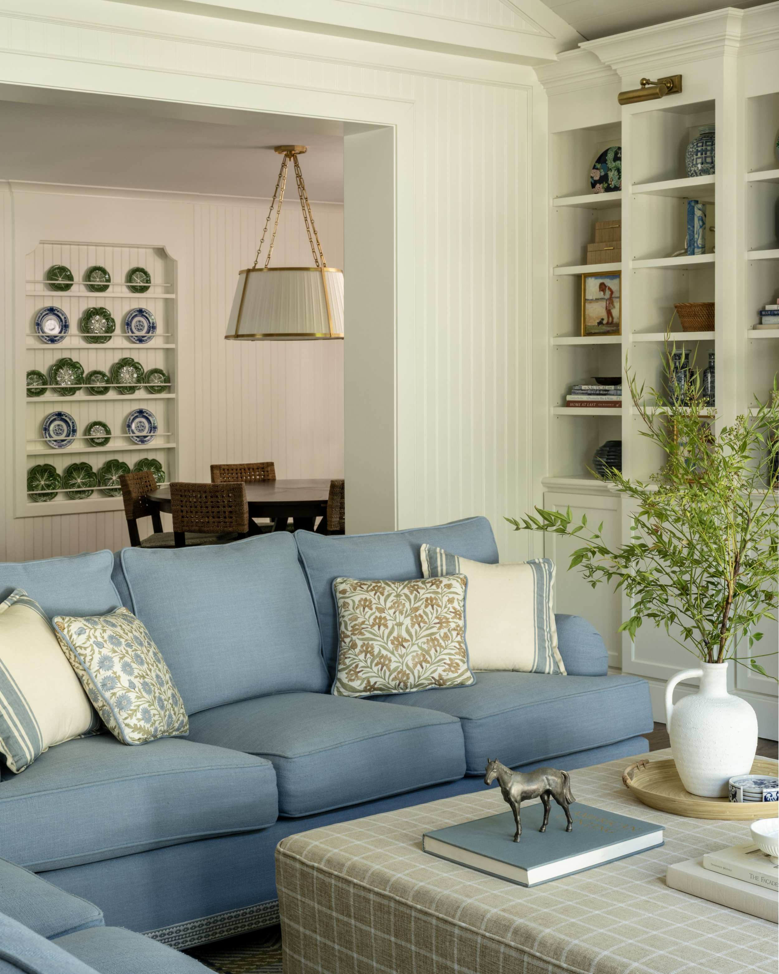 A living room with a light blue sofa and decorative pillows, a white vase with green foliage, and a pedestal table with a horse figurine and books. In the background, a dining area with a wooden table, chairs, a chandelier, and a built-in bookshelf w