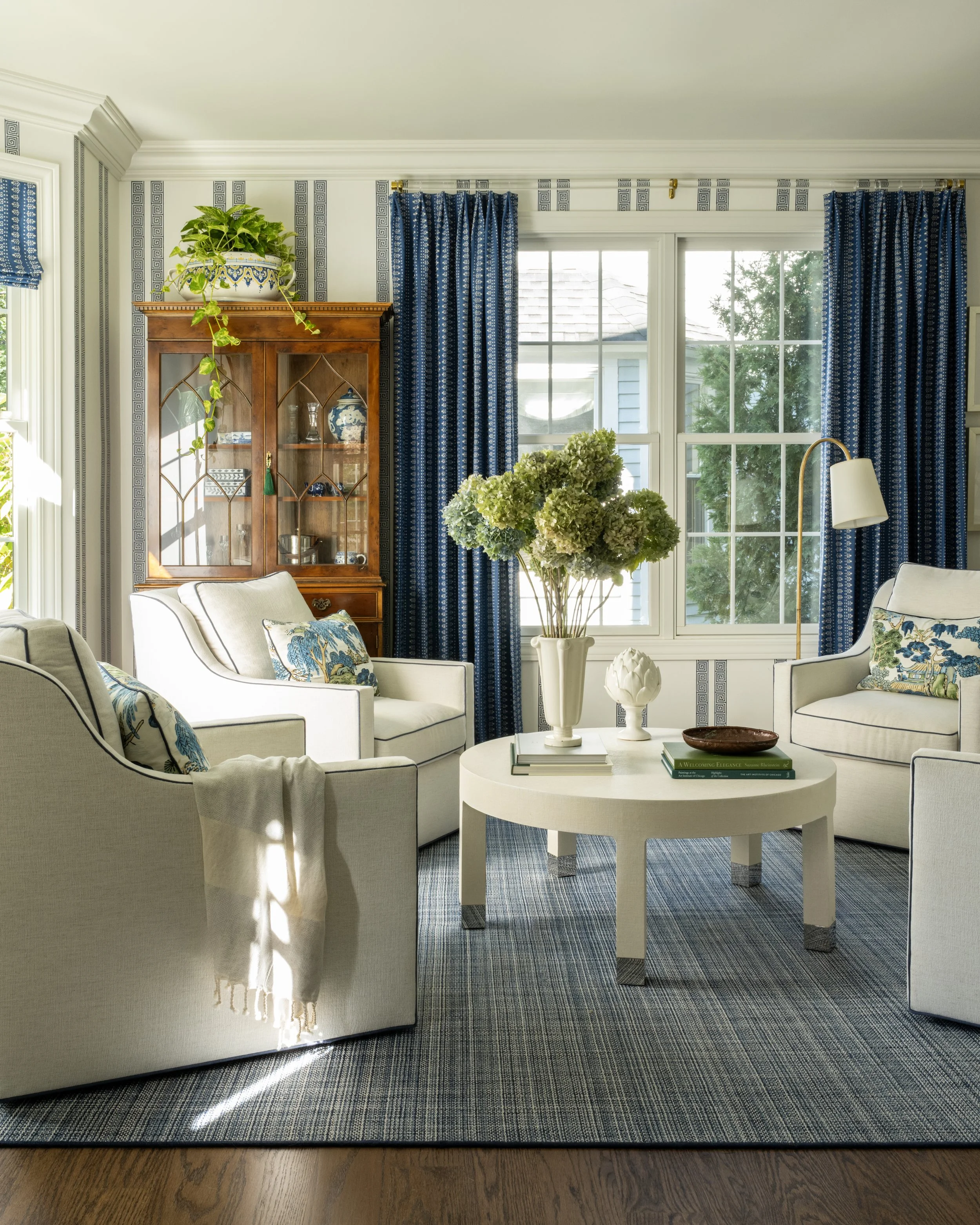 A cozy living room with white armchairs, a round white coffee table, blue patterned curtains, a wooden display cabinet, and large windows letting in natural light.
