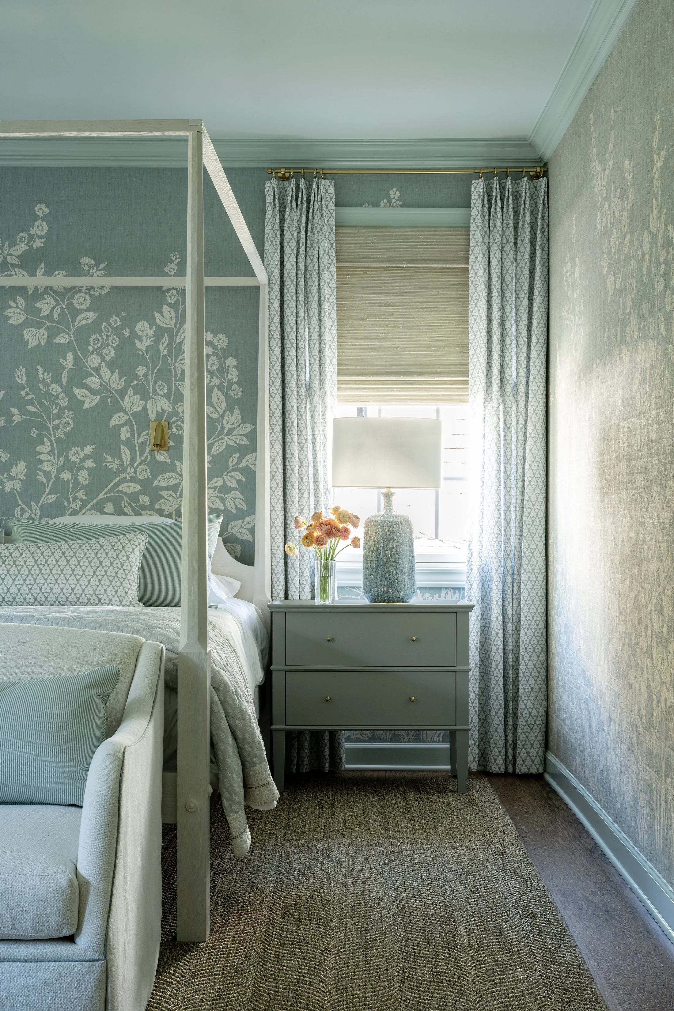 A bedroom corner with a bed, a nightstand with a flower vase and lamp, and a window with curtains and blinds.