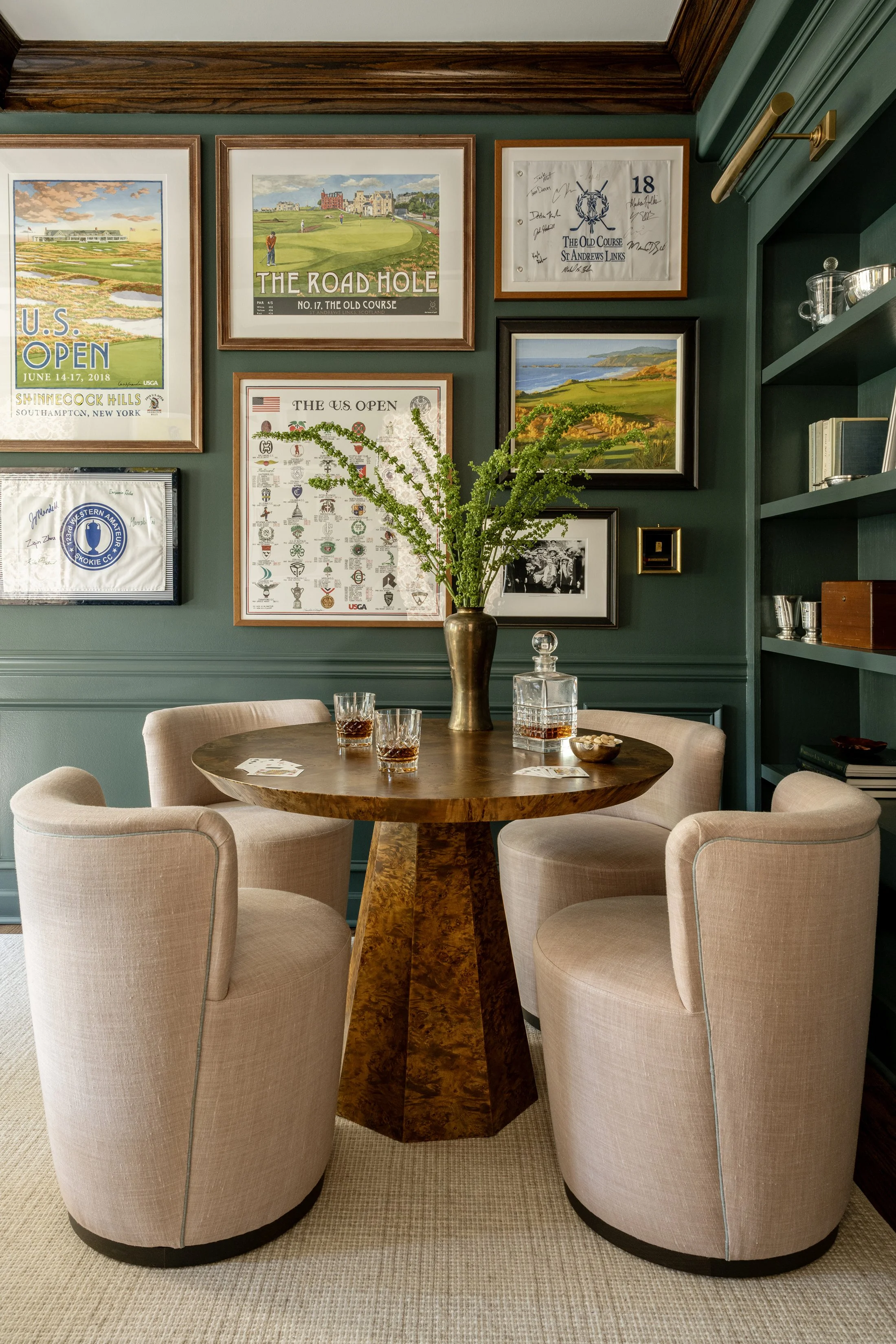 A round wooden table with four beige chairs in a room with dark green walls. The table has a clear decanter, three glasses with drinks, a bowl of snacks, and a tall vase with green leafy branches. The wall behind has framed artwork including golf cou