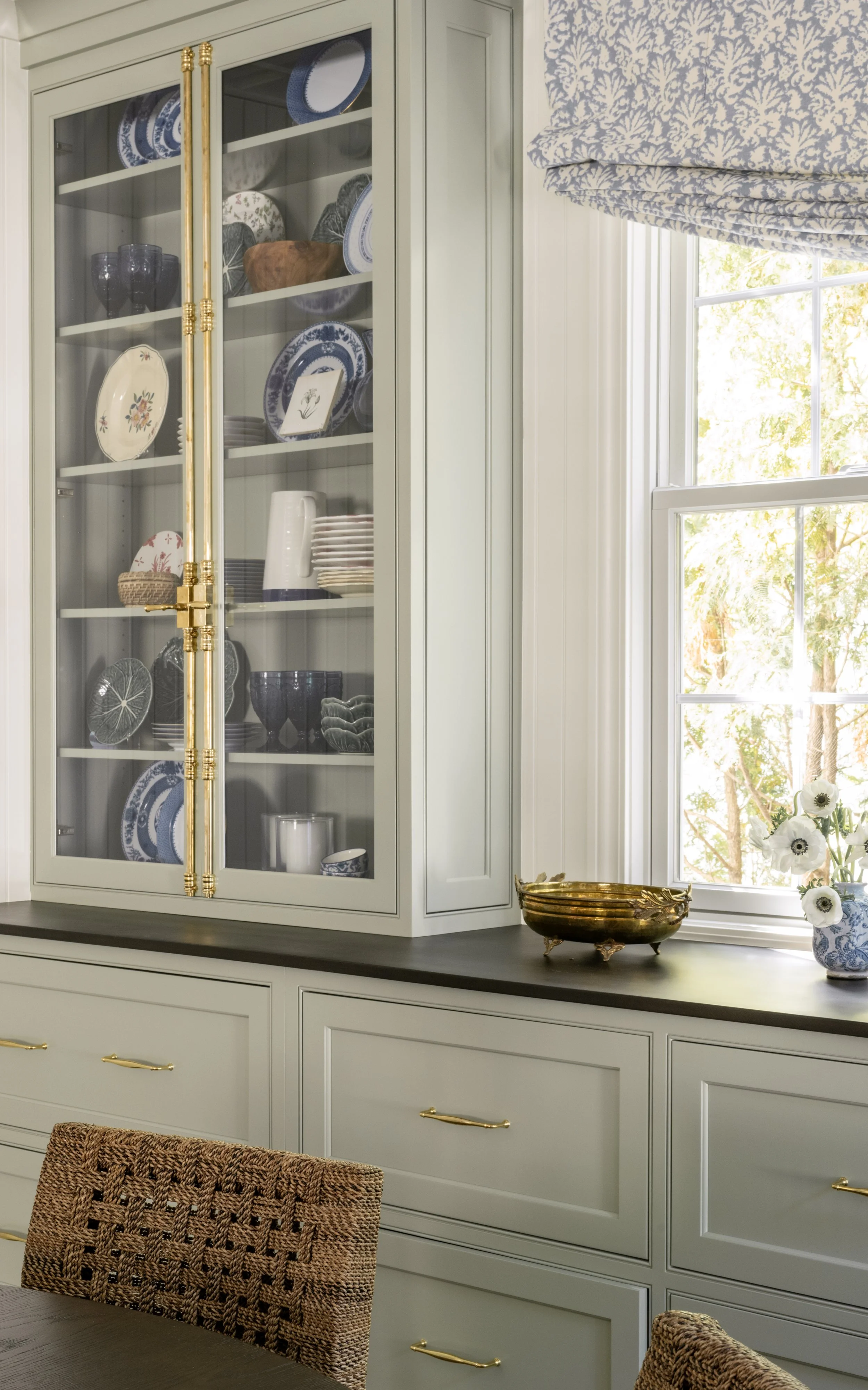 A white china cabinet with glass doors displaying blue and white dinnerware, next to a window with blue patterned curtains, a gold bowl on the dark countertop, and a vase with white flowers.
