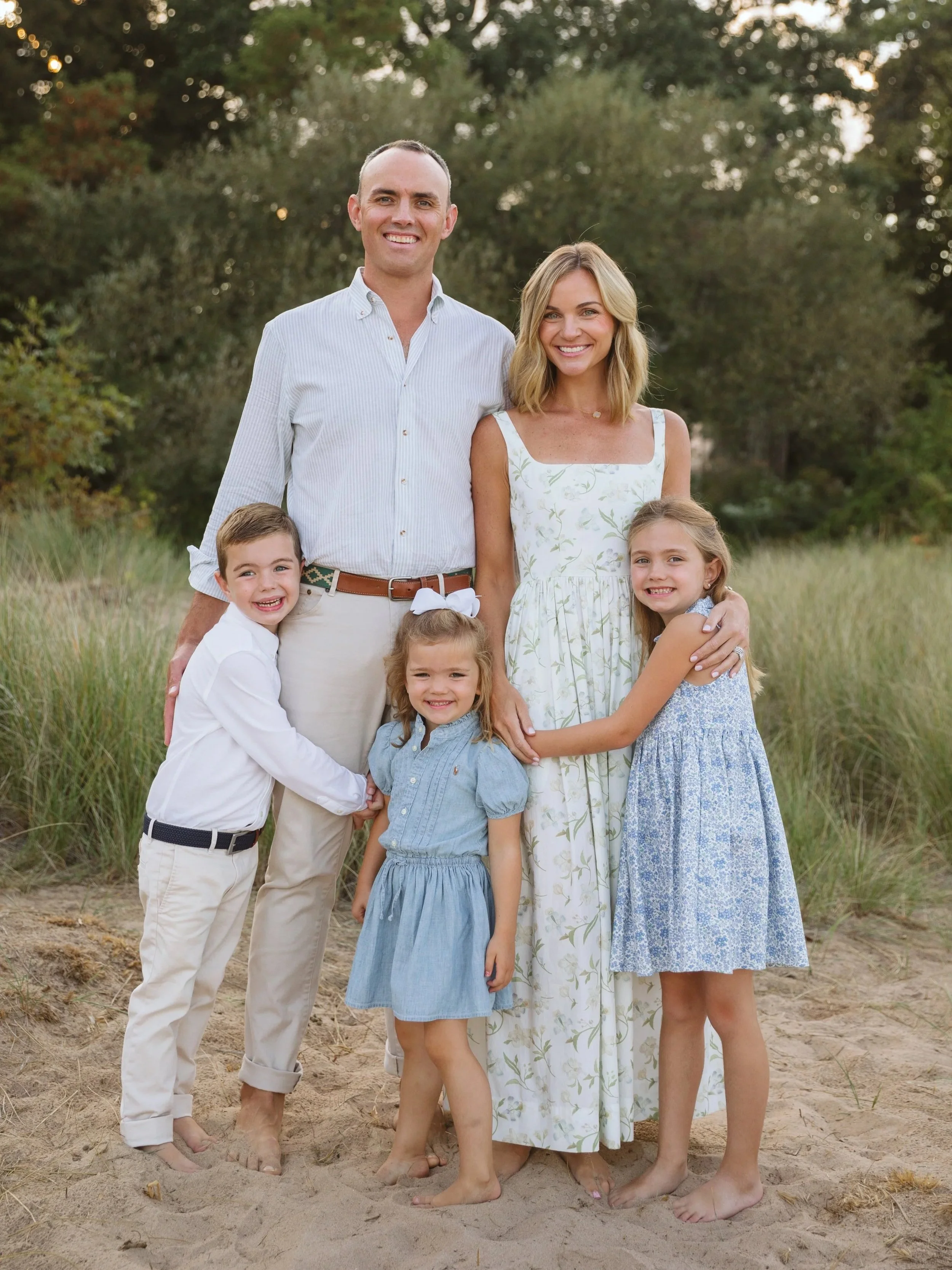 A family of six standing on a sandy beach with greenery in the background. The father and mother are standing in the center, with three young girls and a young boy gathered around them, all smiling and dressed in light-colored, casual clothing.