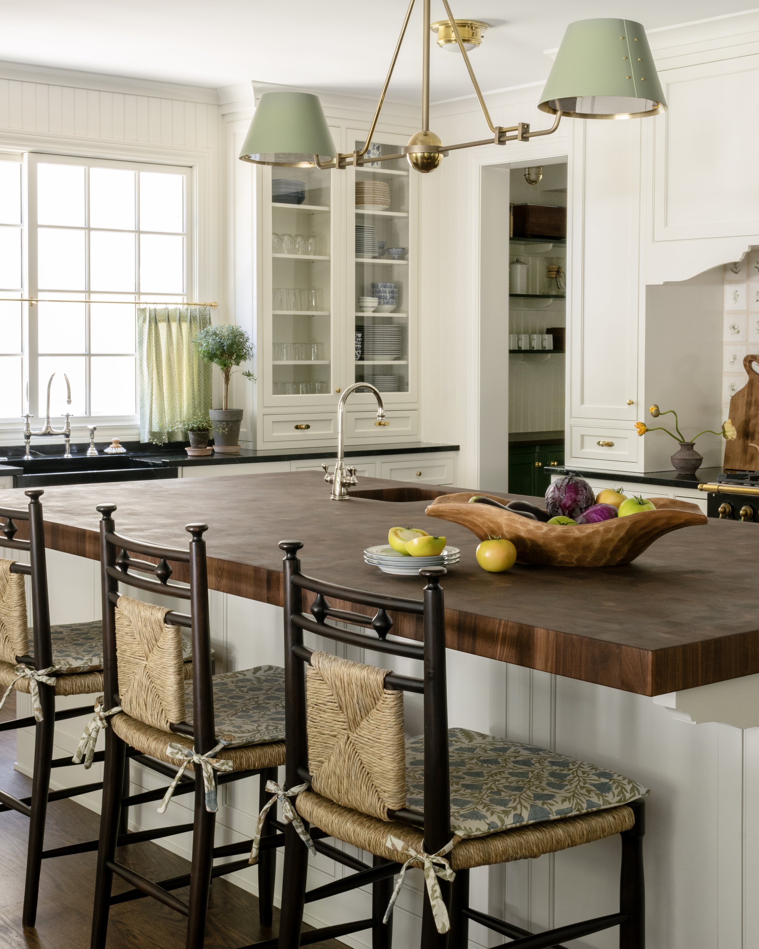A bright kitchen with white cabinets, a wooden island, and bar stools with woven seats. There are green lampshades overhead and a window with green curtains. The island has a wooden bowl with apples and vegetables.