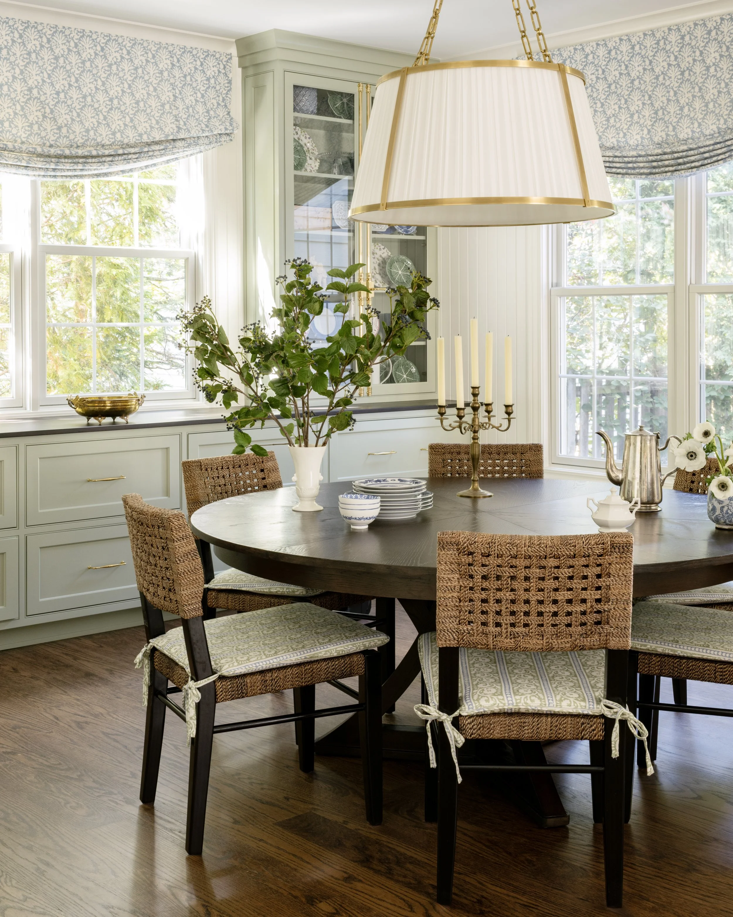 A bright dining room with a round wooden table, six woven chairs with cushioned seats tied with ribbons, a large pendant light hanging overhead, and windows with blue and white patterned curtains. The table is decorated with a white vase of green fol