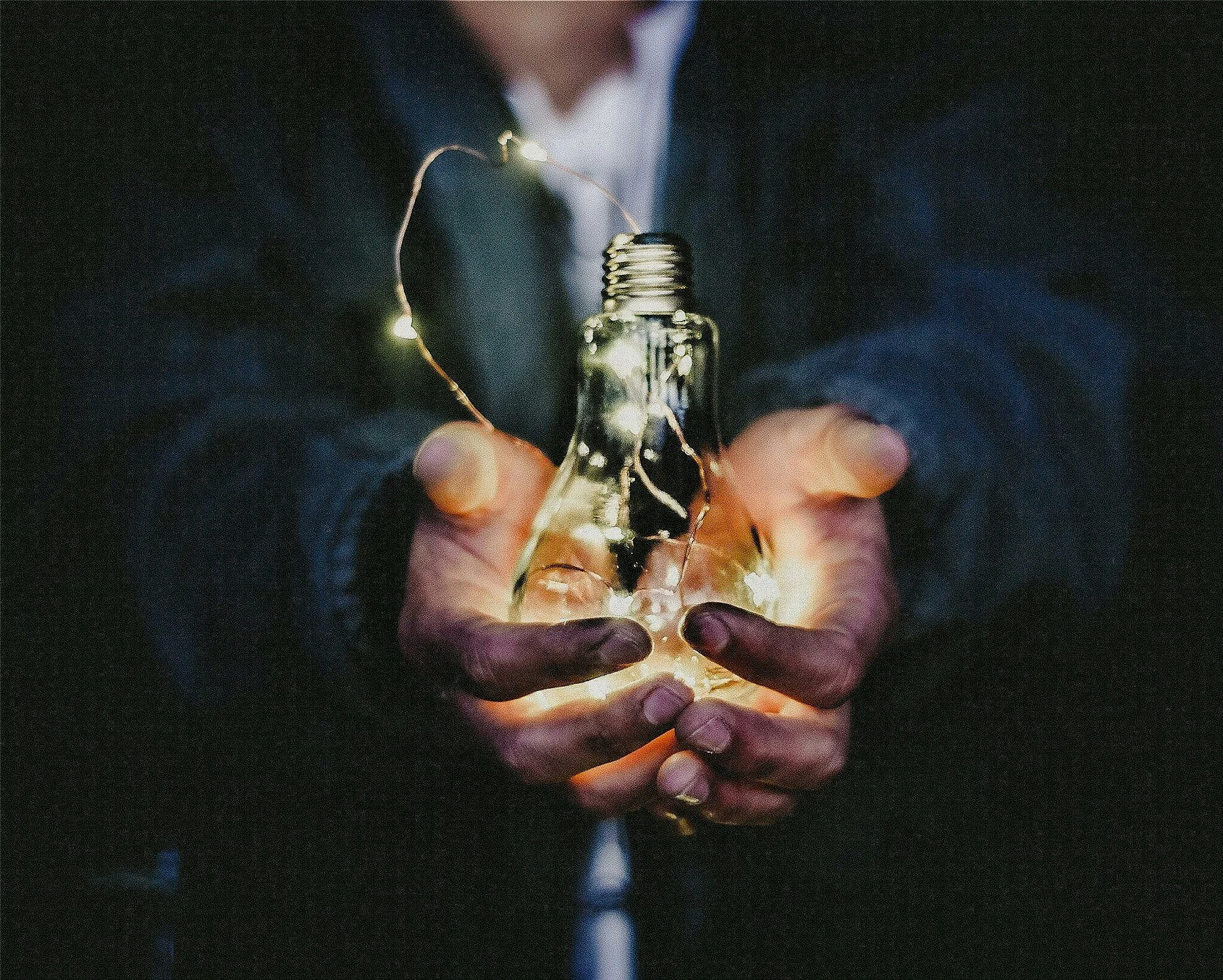 Person holding a glass light bulb with a small string of fairy lights inside it in a dark setting.
