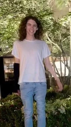 Young woman with curly hair smiling outdoors among trees, wearing a white t-shirt and light jeans.
