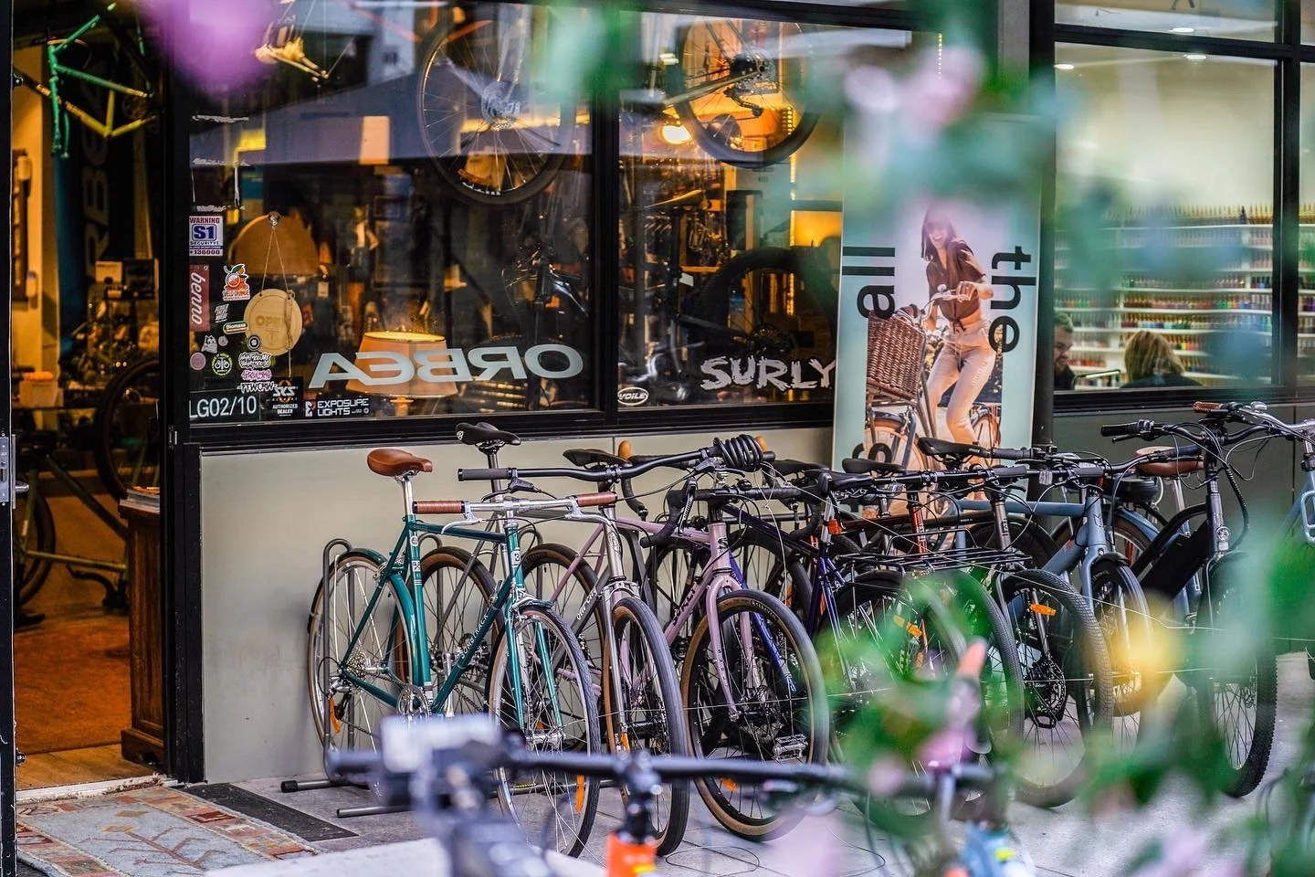 Bicycles parked outside a bike shop with large glass window showing indoor cycling equipment and a poster of a woman with a bike.