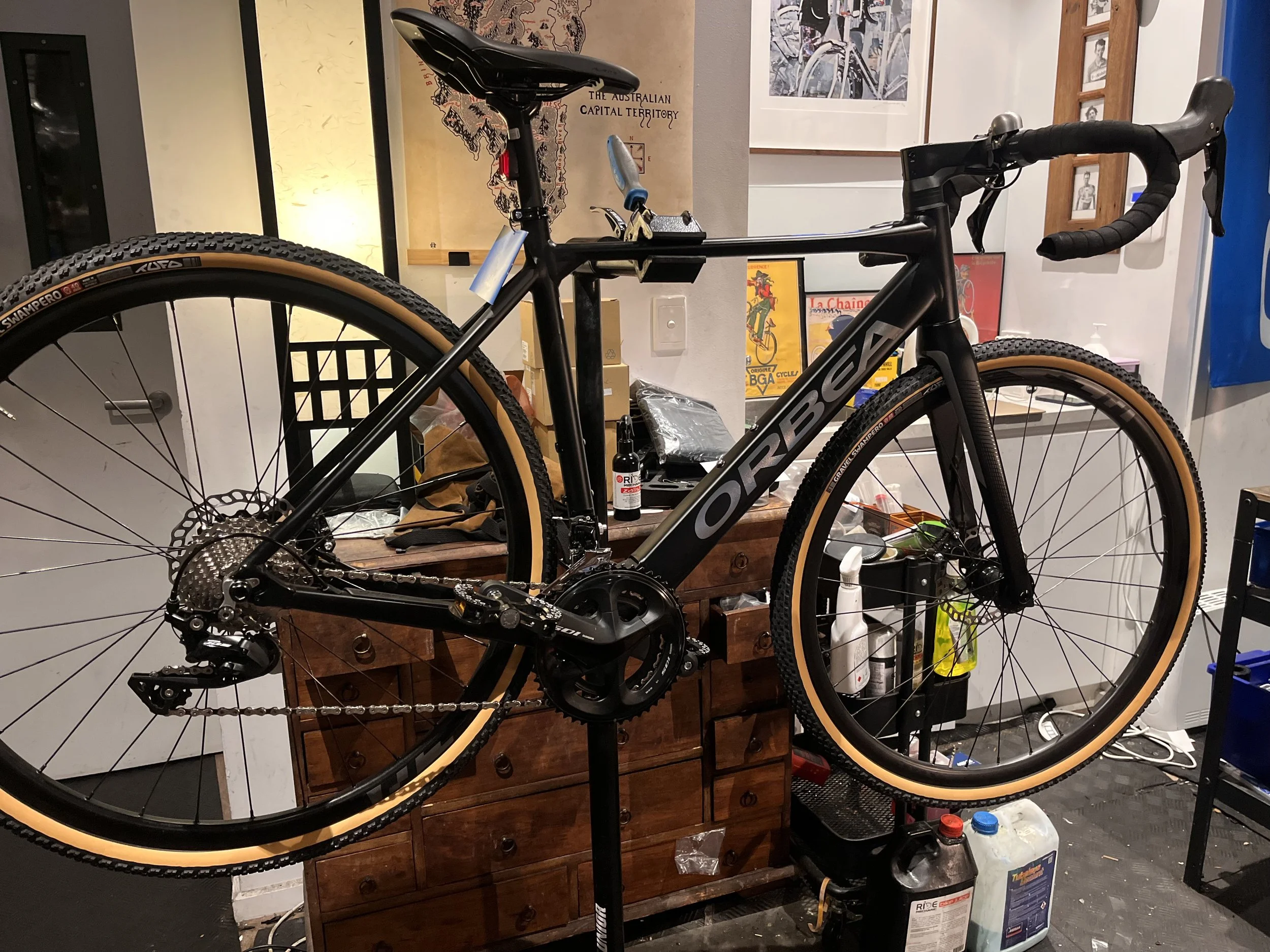 A black ORBEA bicycle mounted on a repair stand in a room, with a wooden dresser and various tools and supplies around it.