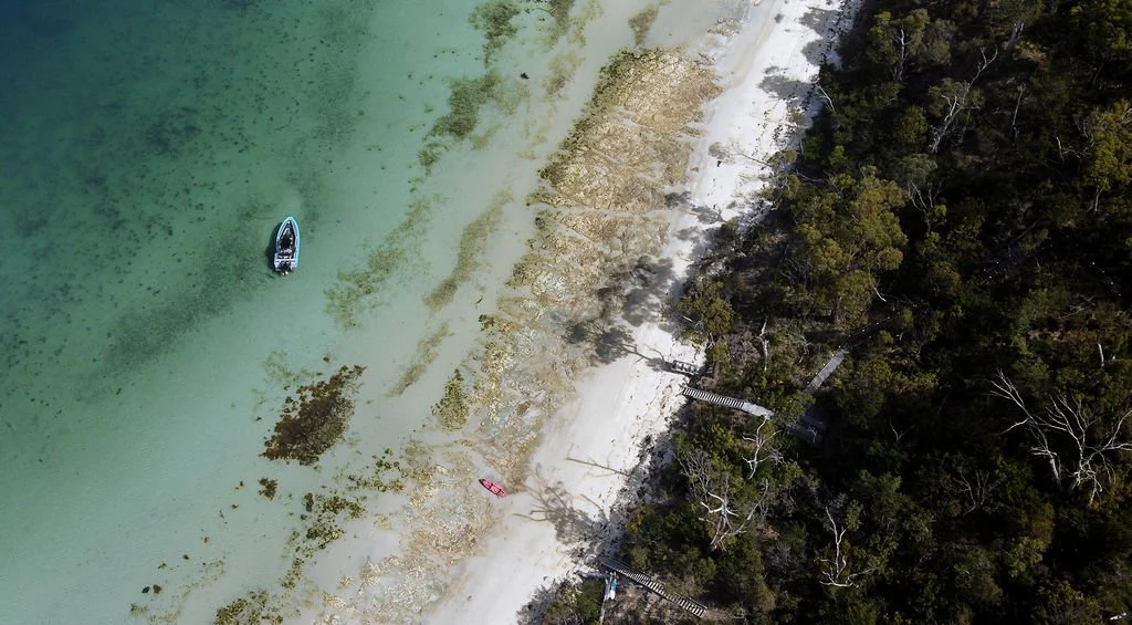 Aerial view of a beach with clear water, a small boat near the shoreline, and surrounded by dense green forest.