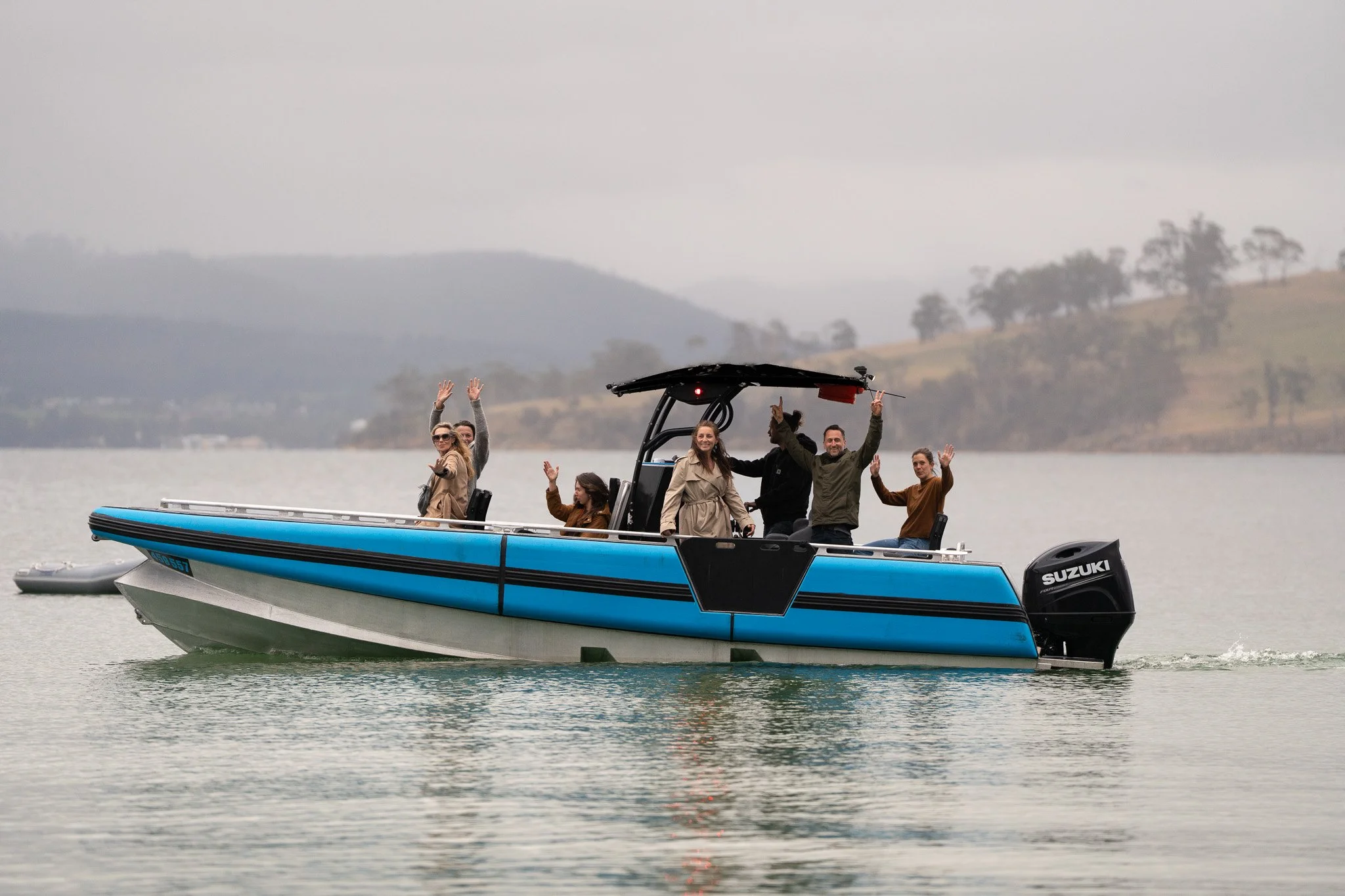 Group of people on a blue boat waving and smiling on a lake with hills in the background.