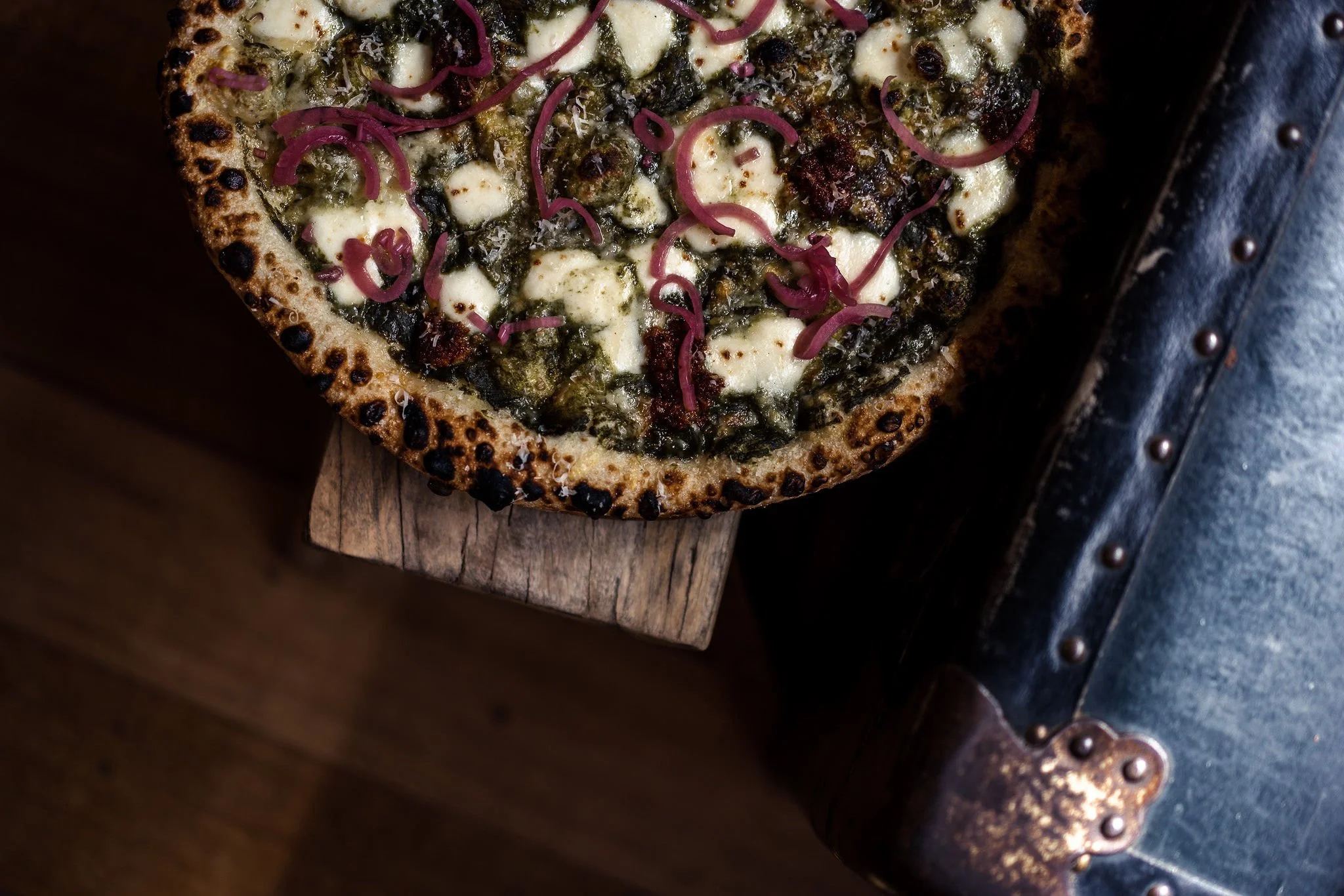 A slice of pizza with a dark crust, topped with cheese, spinach, and pickled red onions, placed on a wooden cutting board.