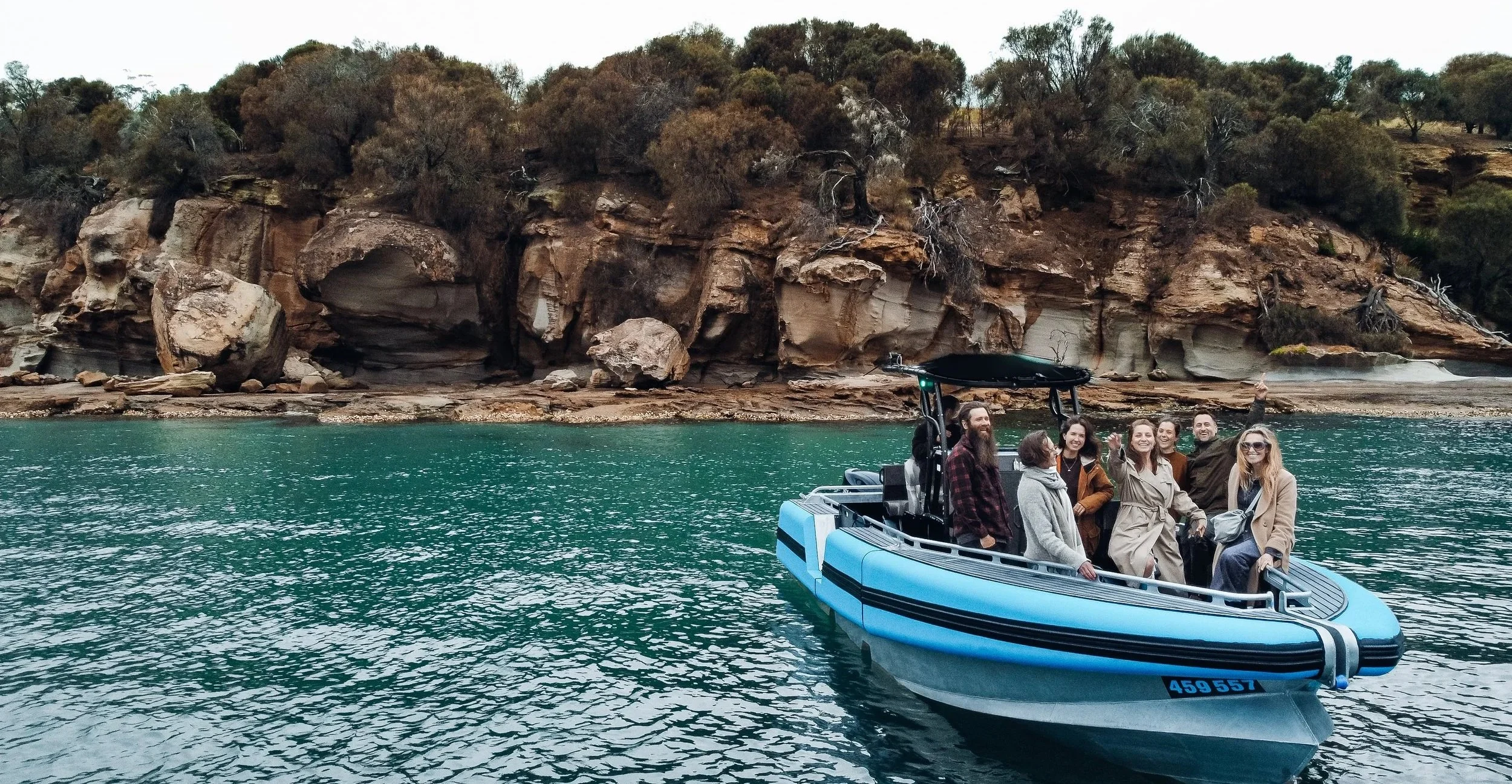 A group of people on a blue and black motorboat on calm water with rocky Bruny Island cliffs and sparse trees in the background.