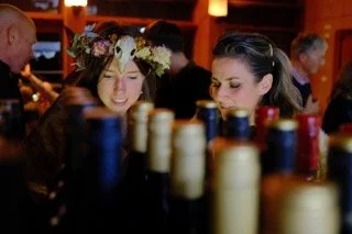 Two women sitting at a bar or restaurant table, surrounded by bottles, one wearing a flower crown.