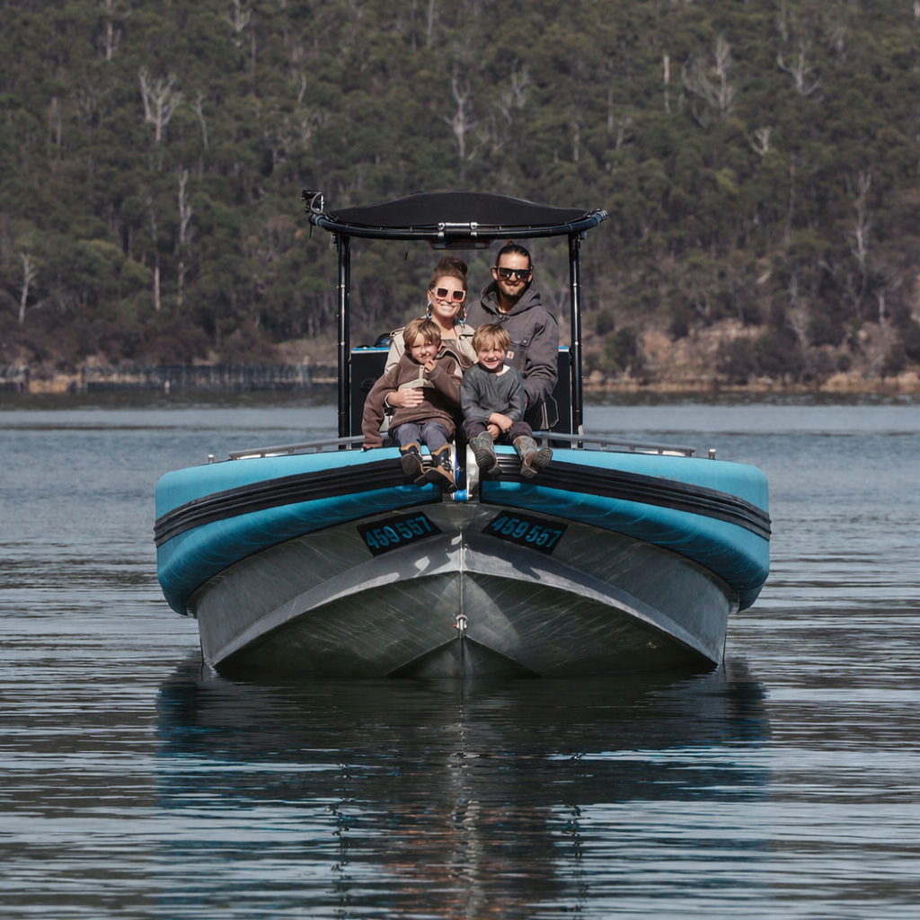 A family of four sitting on the bow of a boat on a calm lake, with trees and hills in the background.