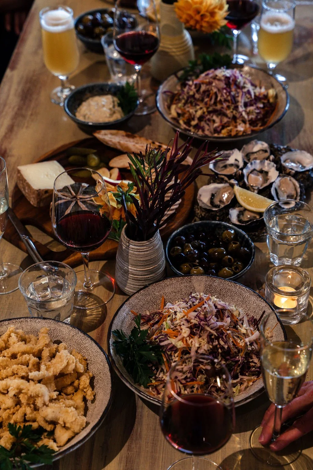 A table set for a meal with various dishes including salads, oysters, cheese, olives, and drinks like red wine, beer, and water, decorated with flowers and candles.