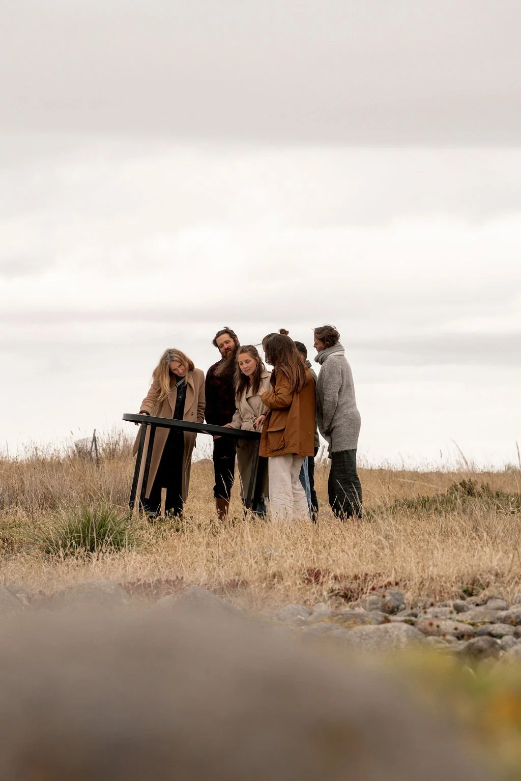 Group of people standing outdoors on a grassy field, looking at and interacting with a small table or device, with overcast sky in the background.