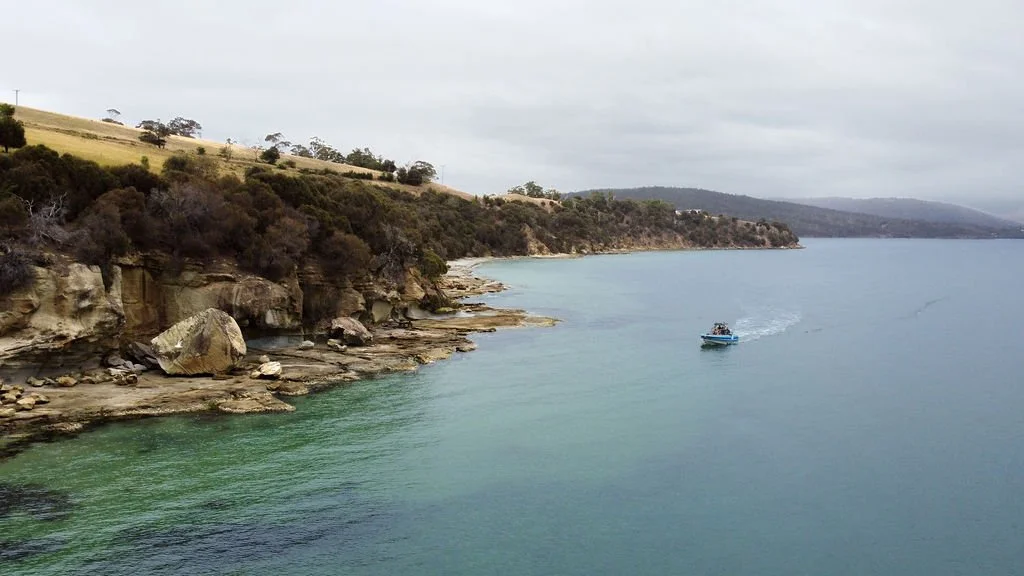 A boat sailing in a calm body of water near rocky shoreline with green hills in the background under an overcast sky.