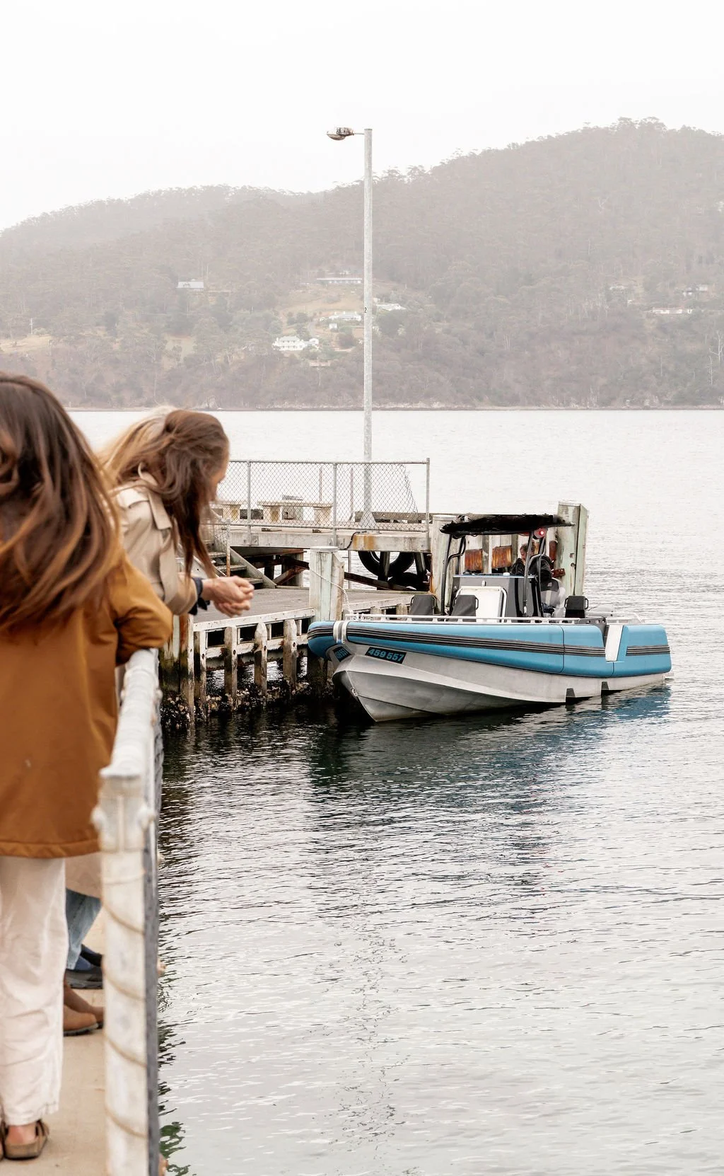 People standing on a dock looking at a blue and white boat docked at the pier with a large body of water and hills in the background.
