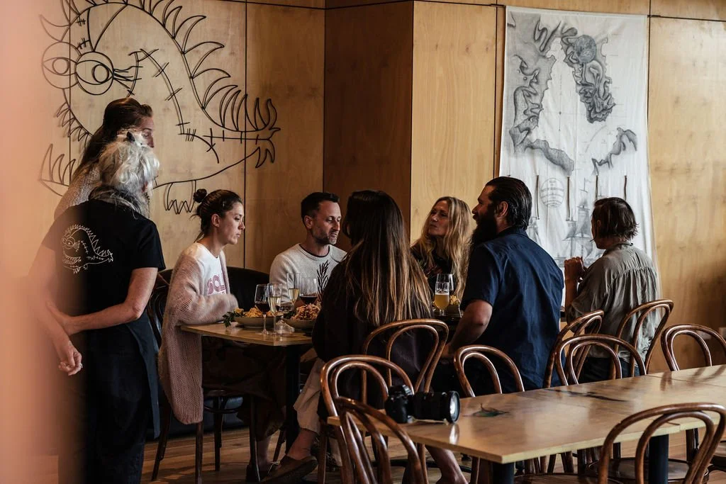 Group of people sitting at a restaurant table, engaging in conversation, with drinks and food. Two women are standing nearby, one with gray hair and one with a bun. The decor includes a large line-art drawing of a dragon and a black-and-white abstrac