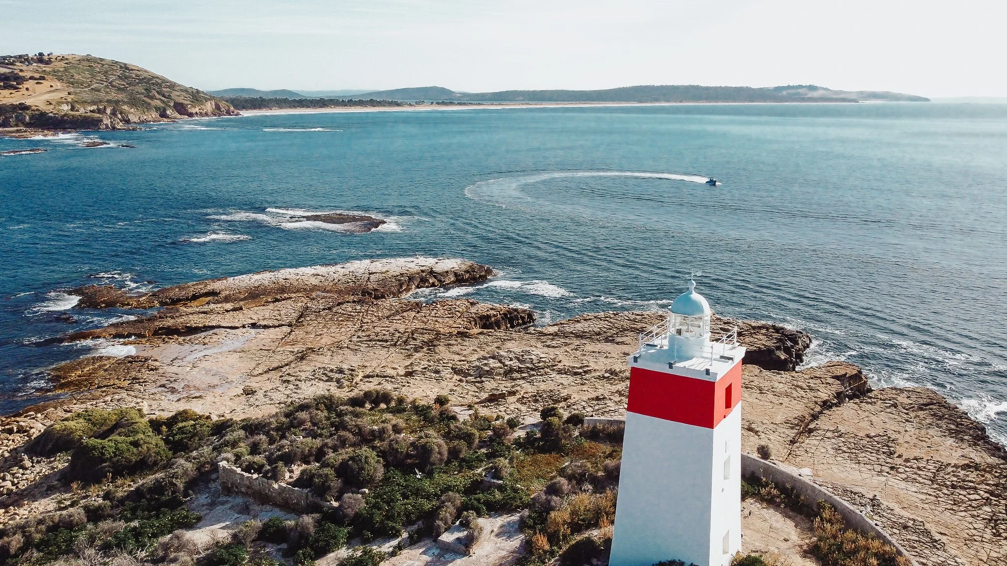 A lighthouse on a rocky coastline near the ocean, with a boat speeding across the water in the distance, and hills in the background under a clear sky.