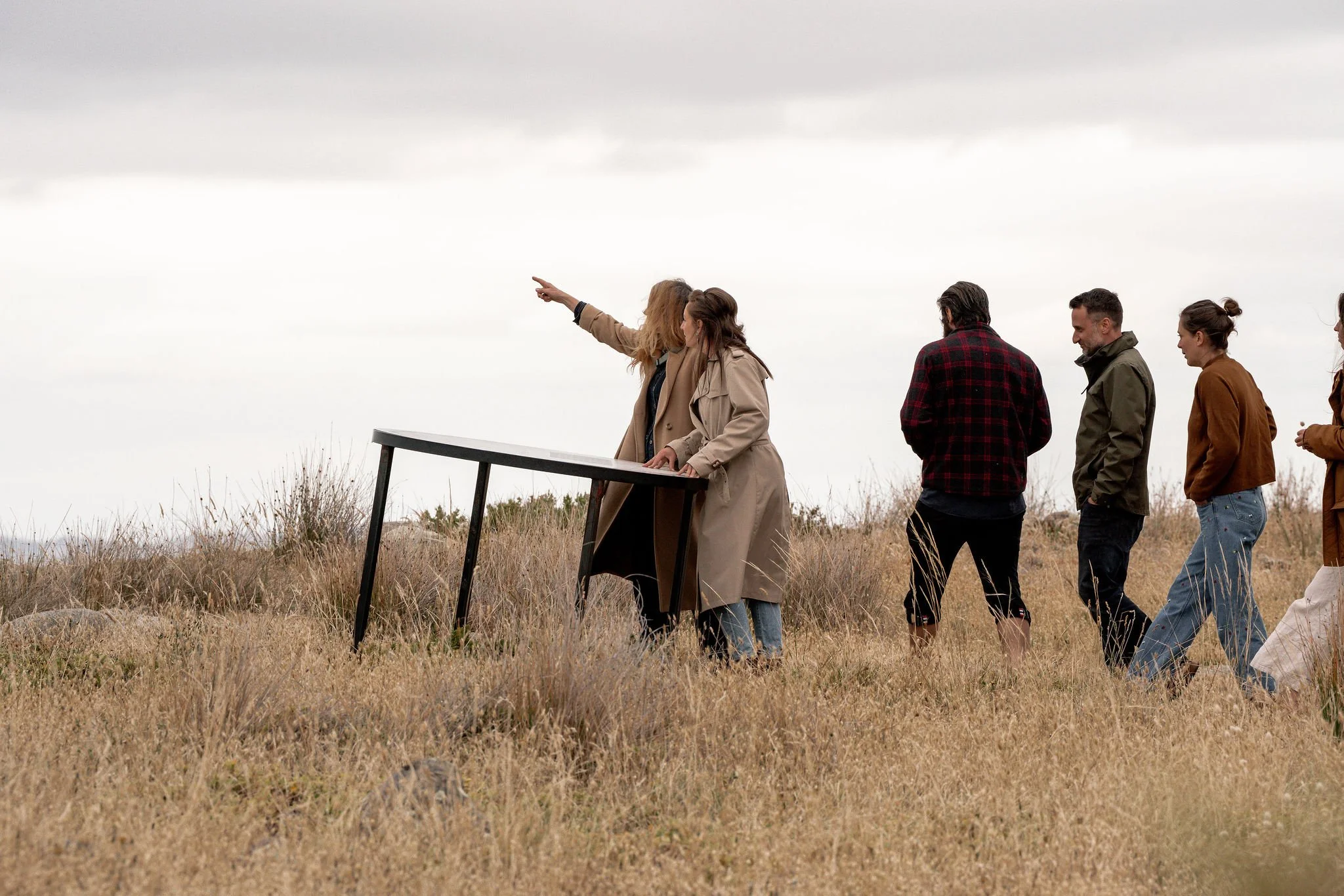 Group of people outdoors, standing in a line on a grassy field, with two women at the front, one pointing and the other looking at the scenery, under an overcast sky.
