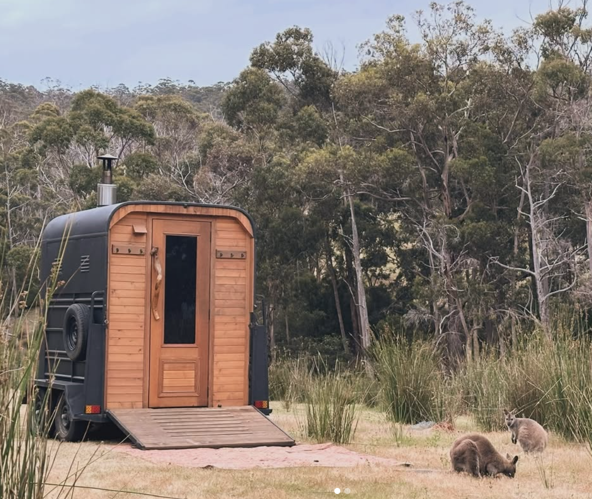 A small black Sauna trailer with wooden door and ramp set up in a grassy field surrounded by trees, with kangaroos resting on the grass nearby.