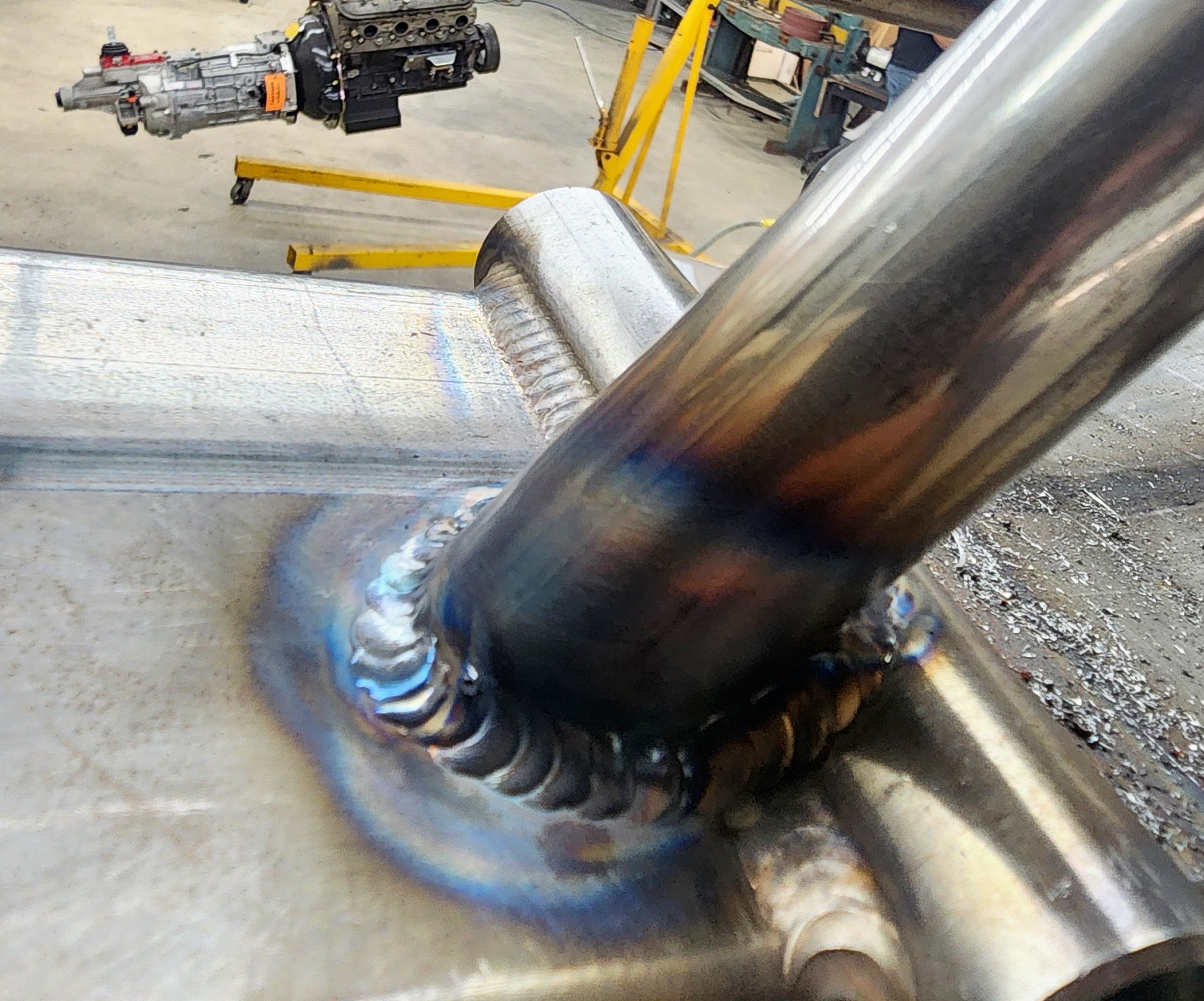 Close-up of a metal weld joint on a pipe, with a car engine and transmission in the background, inside a workshop.