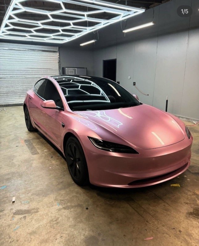 A pink Tesla Model 3 parked in an indoor garage with geometric LED lighting on the ceiling.