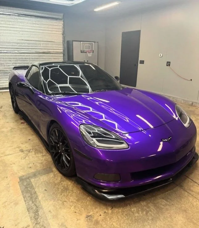 A purple Chevrolet Corvette sports car with a custom front splitter, parked inside a garage with concrete floor and gray walls.