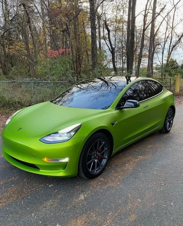 Green Tesla Model 3 parked on a street with trees in the background.