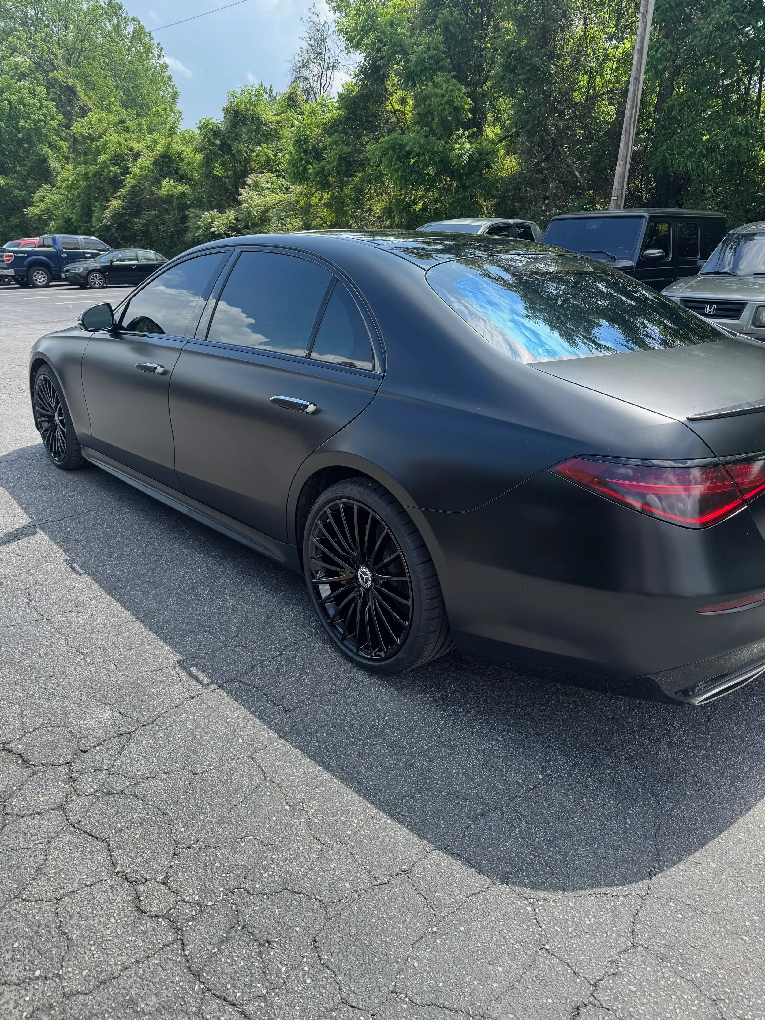 A black luxury sedan with tinted windows parked on a cracked asphalt lot, with trees and other vehicles in the background.