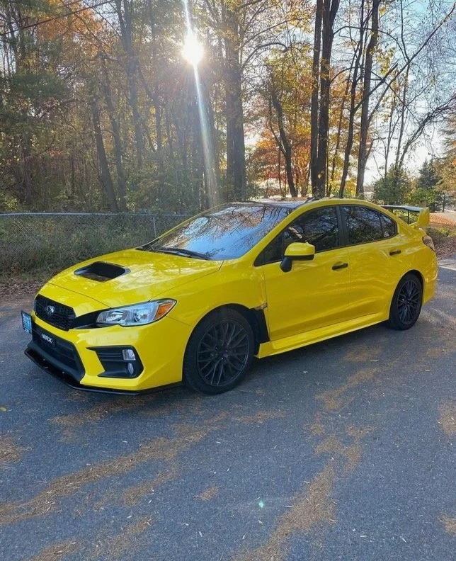 Yellow sports car with black wheels parked on a paved road near trees with fall foliage, with sunlight shining through the trees.