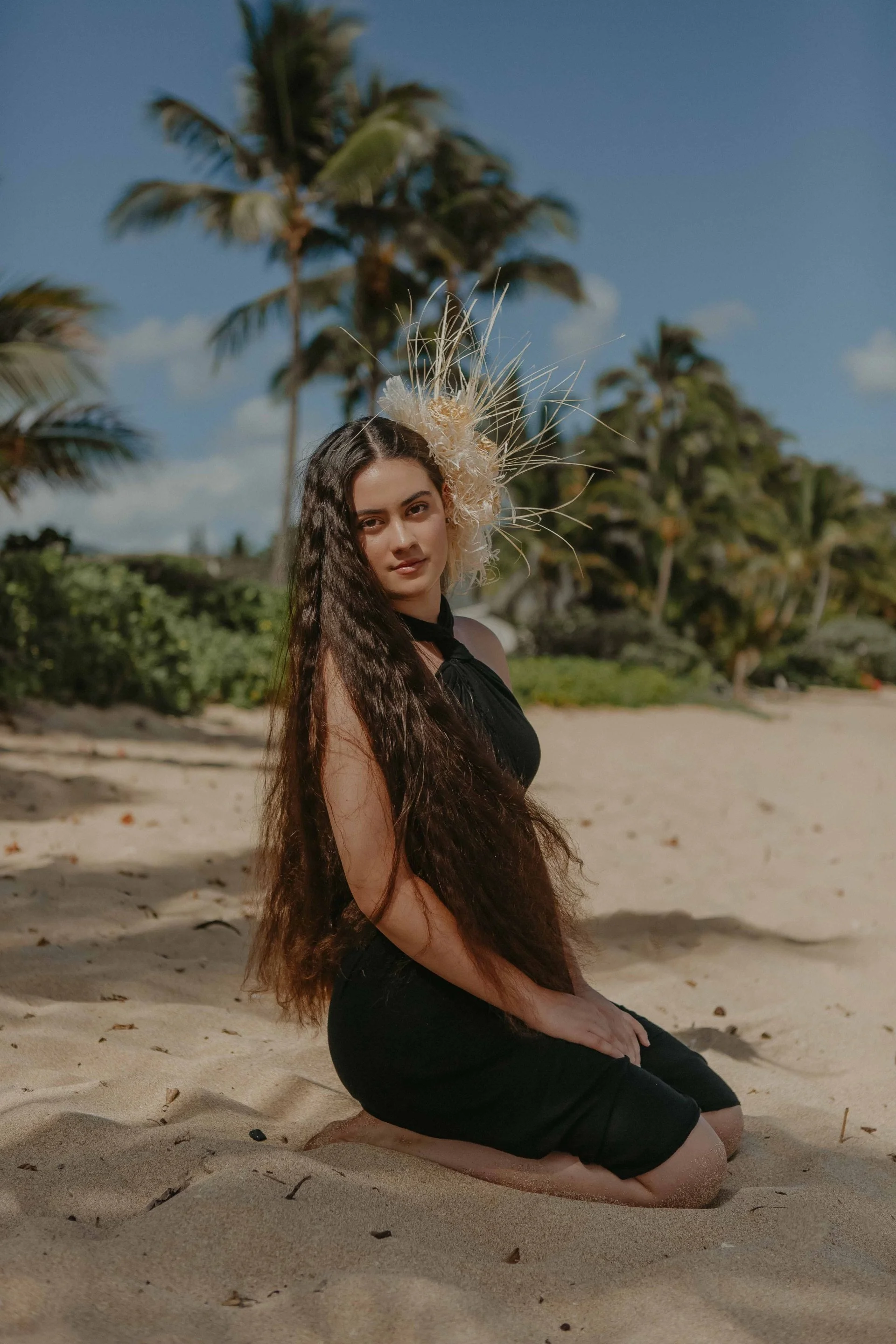 A woman with long dark hair, sitting on her knees on a sandy beach, wearing a black pareu with a decorative with woven headpiece, and palms trees in the background.