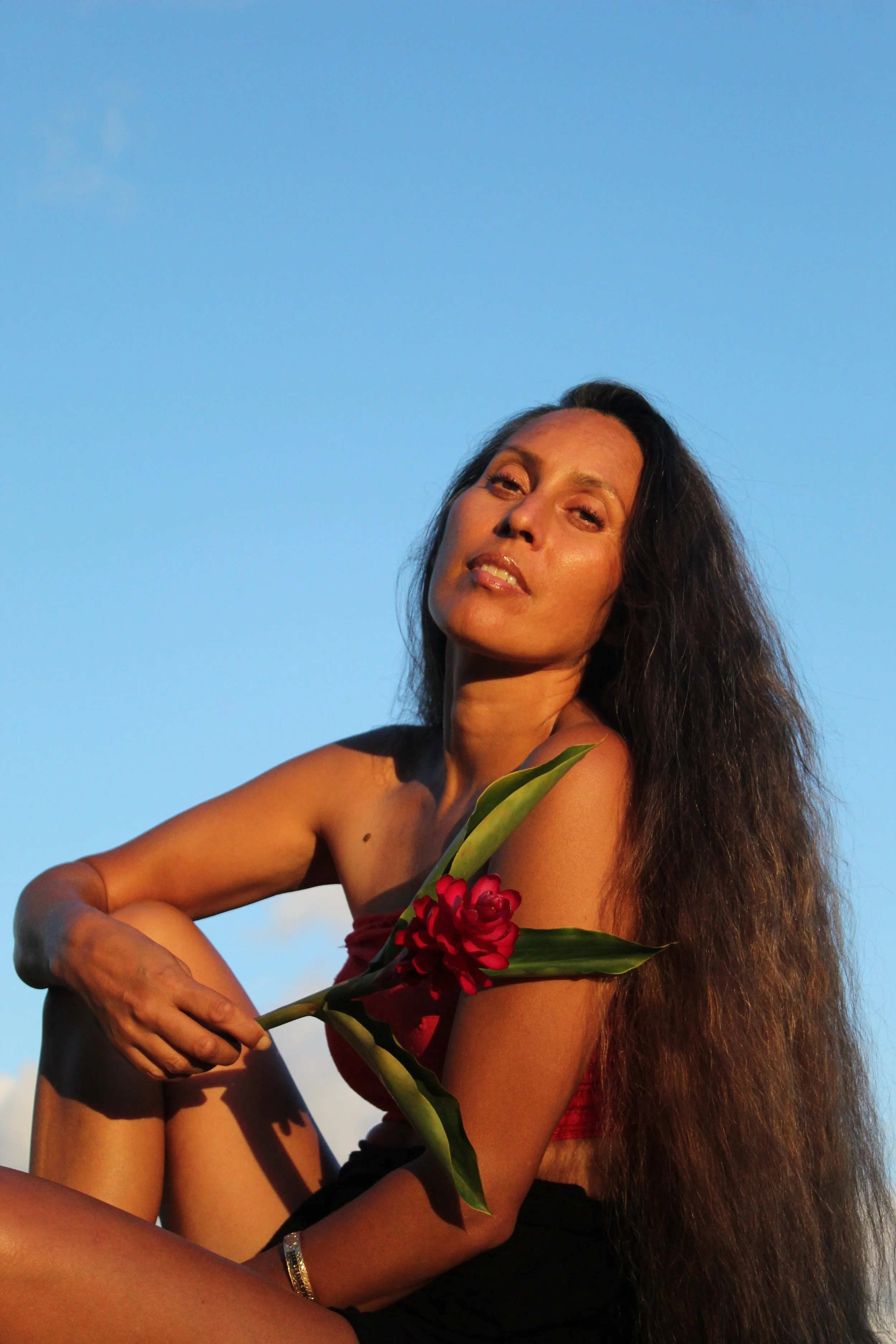 A woman with long dark hair sitting outdoors against a clear blue sky, holding a red flower and a green leaf, with a serene expression.