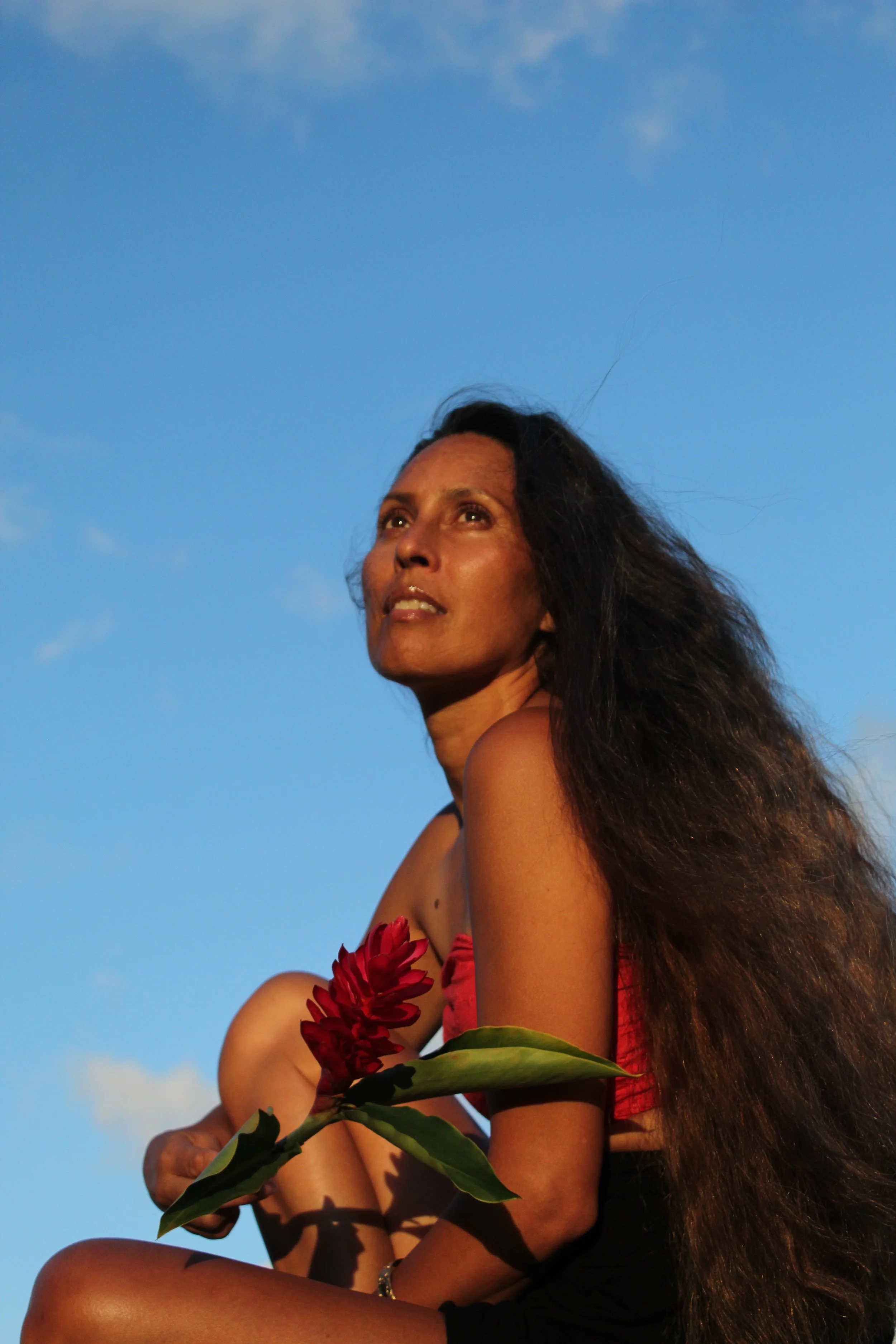 A woman with long dark hair, tan skin, and a red flower in her hand, sitting outdoors with a blue sky background.
