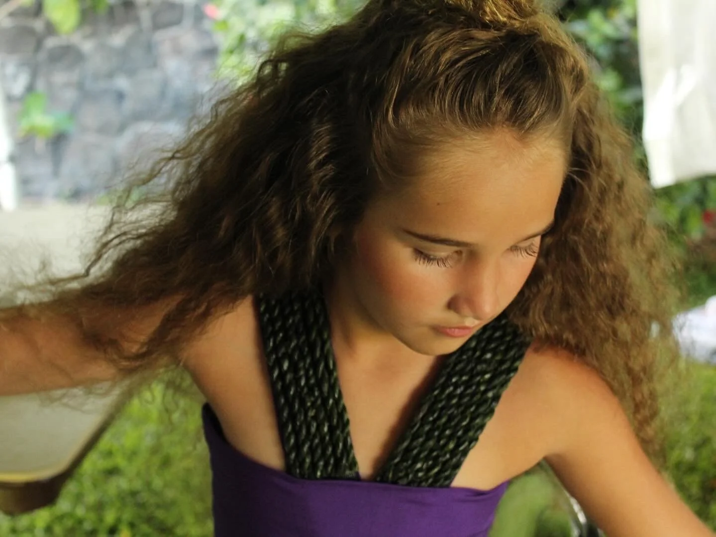 Close-up of a young woman with curly hair looking down, outdoors surrounded by greenery and getting ready for a Tahitian dance competition.