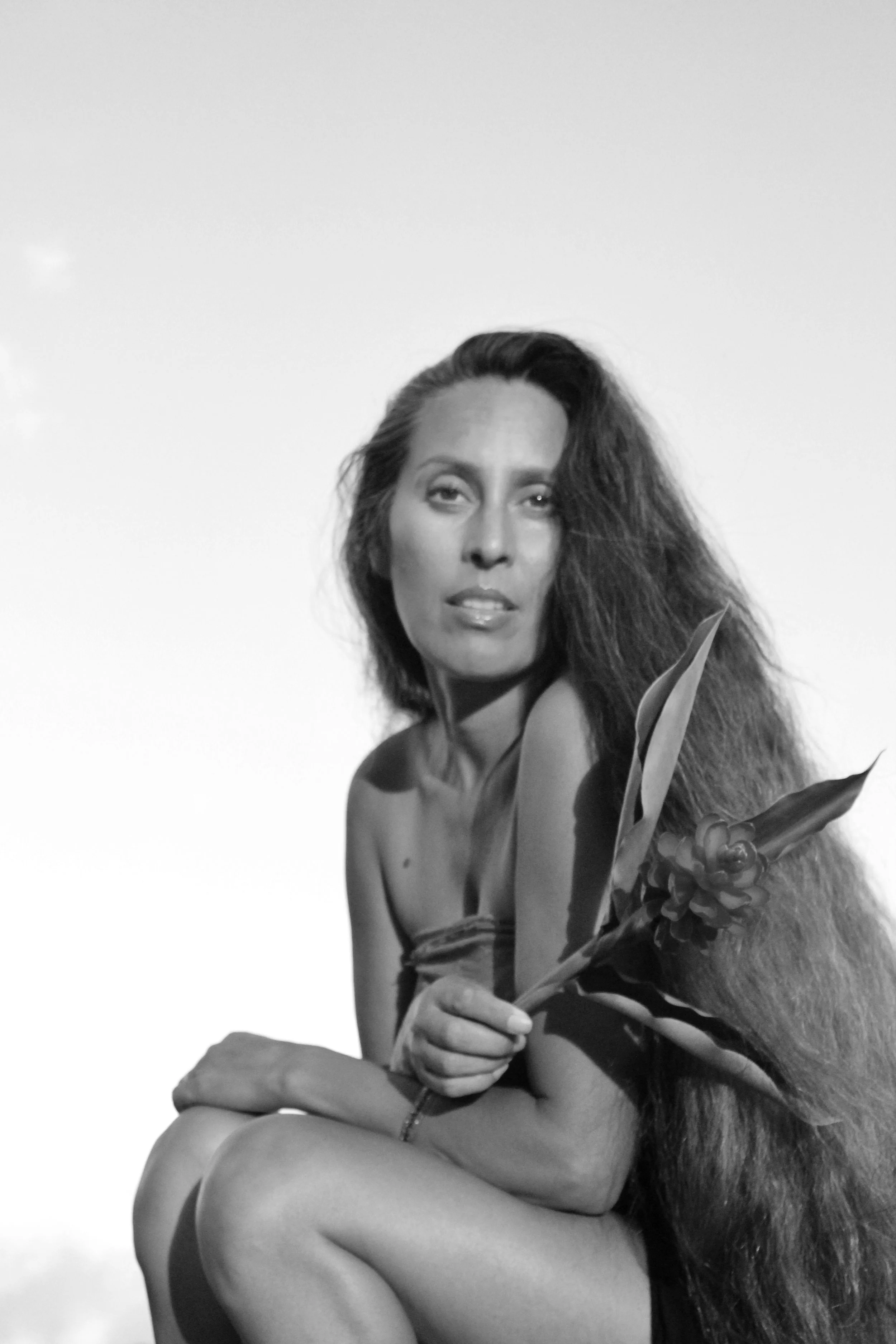 Black and white photo of a woman with long hair, seated with her arms crossed, holding a flower, looking at the camera.