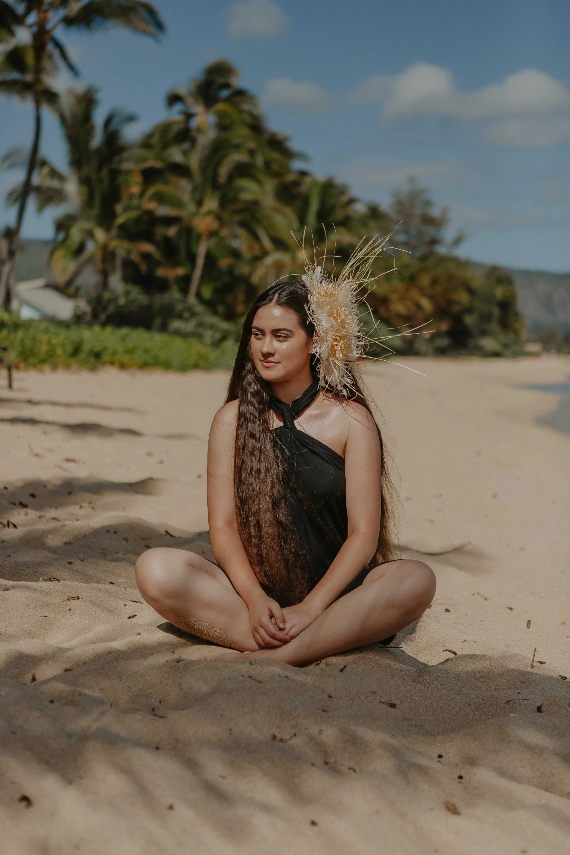 A woman with long dark hair sitting cross-legged on a sandy beach, wearing a black pareu and a woven headpiece. Palm trees and blue sky with a few clouds are in the background.