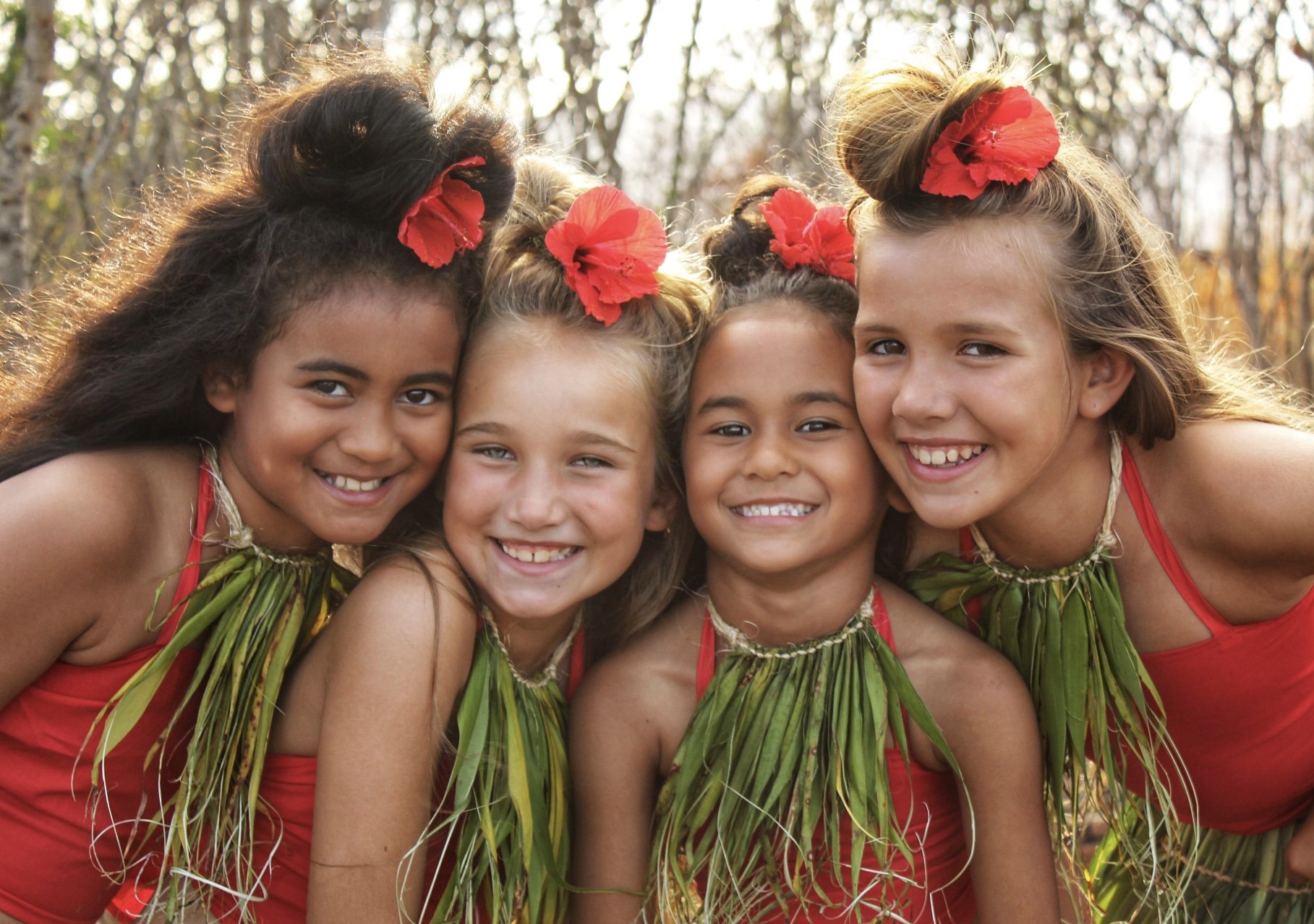 Five smiling girls with flowers in their hair and green and red traditional outfits, united close together outdoors with trees and sunlight in the background.